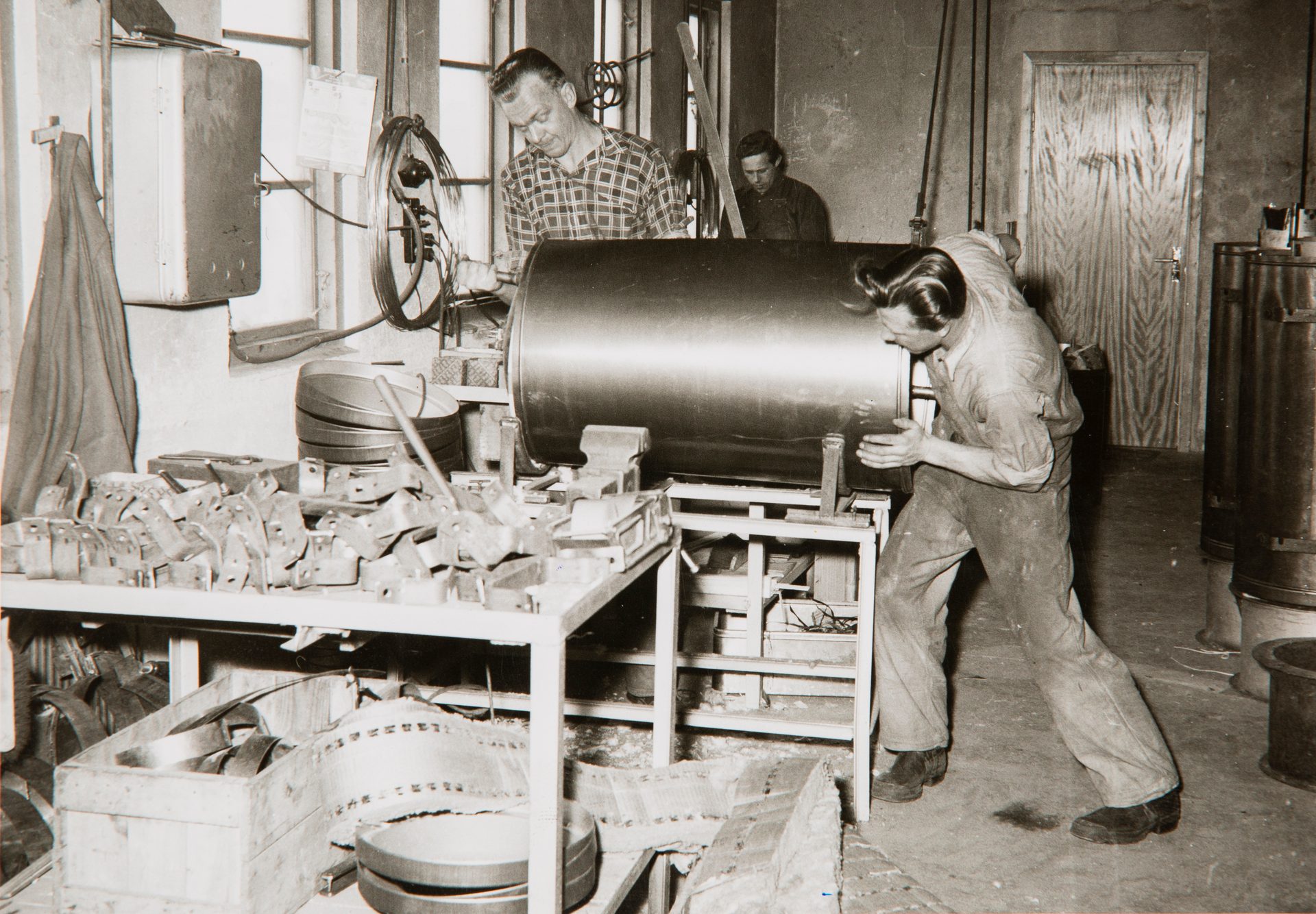 Three men work in a vintage workshop, two focused on a large metal cylinder, one in background.