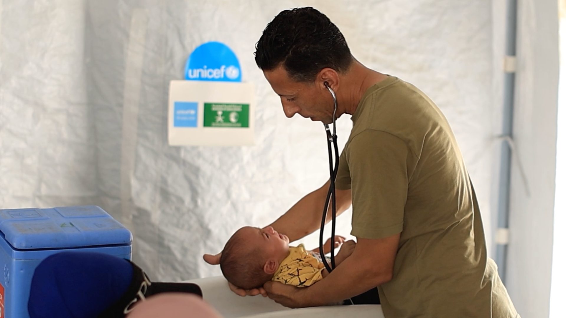 A doctor uses a stethoscope to examine a baby on a white examination surface, with a UNICEF logo in the background.