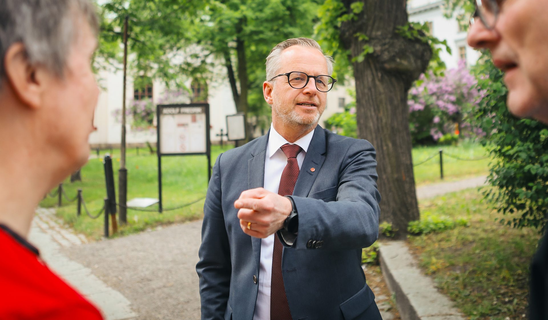 A man in a suit speaks and gestures to two people in an outdoor setting.