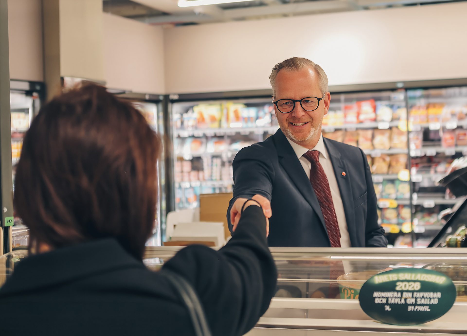 A smiling man in a suit shakes hands with a woman (seen from behind) in a grocery store aisle.