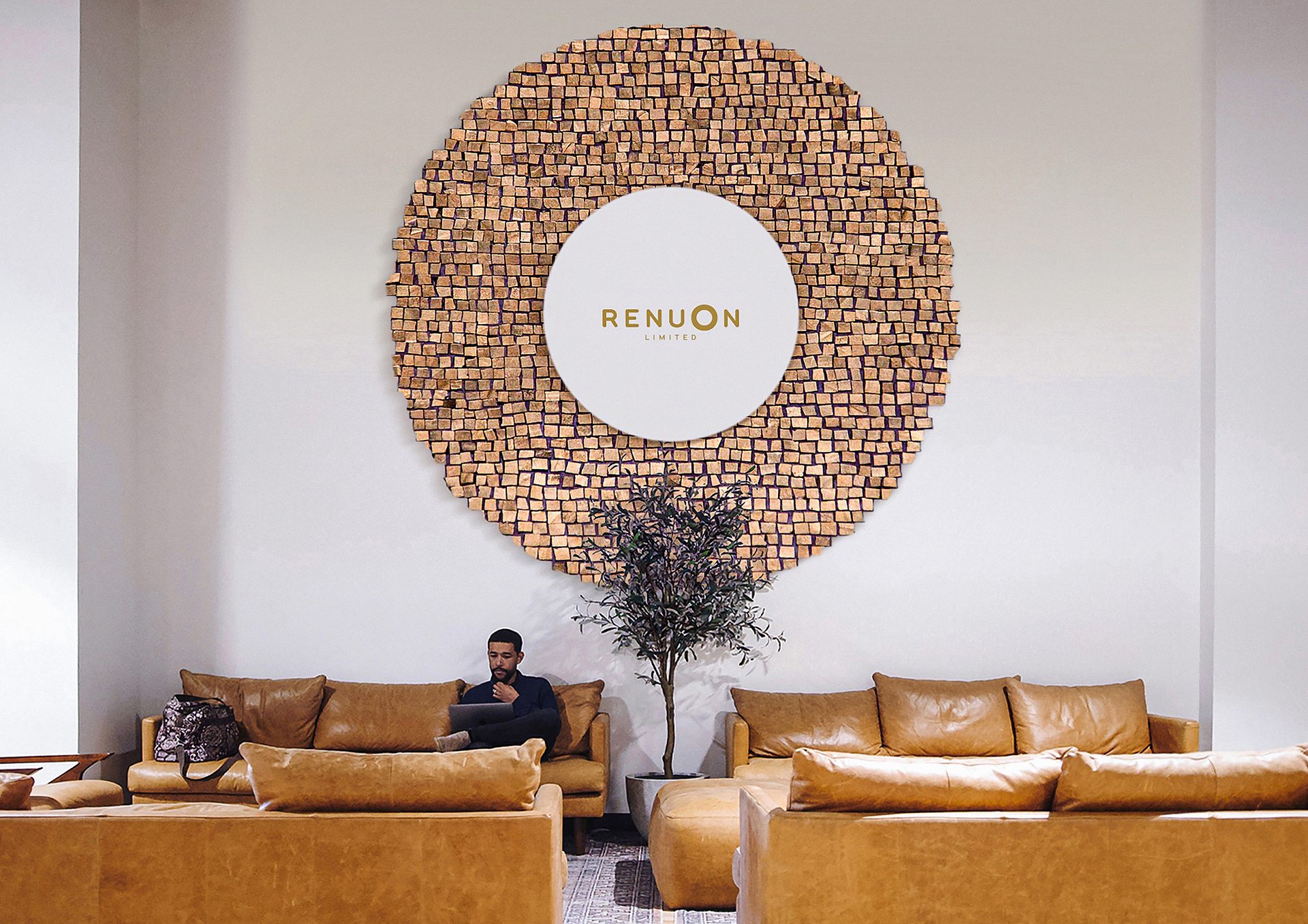 Man on leather couch with laptop, under a large circular wooden Renuon Limited wall art.