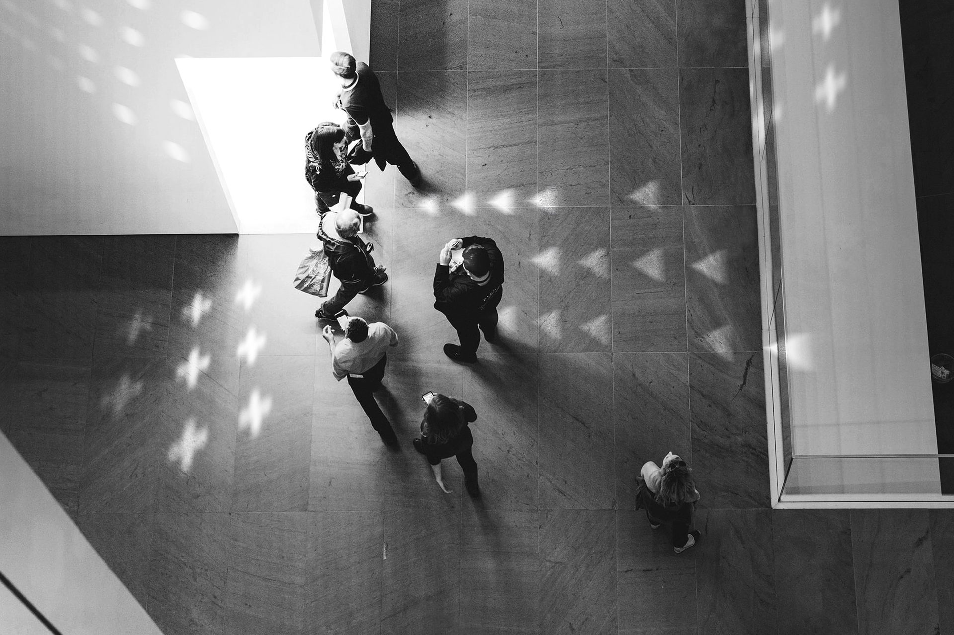 Overhead B&W photo of people on a tiled floor with geometric light patterns.