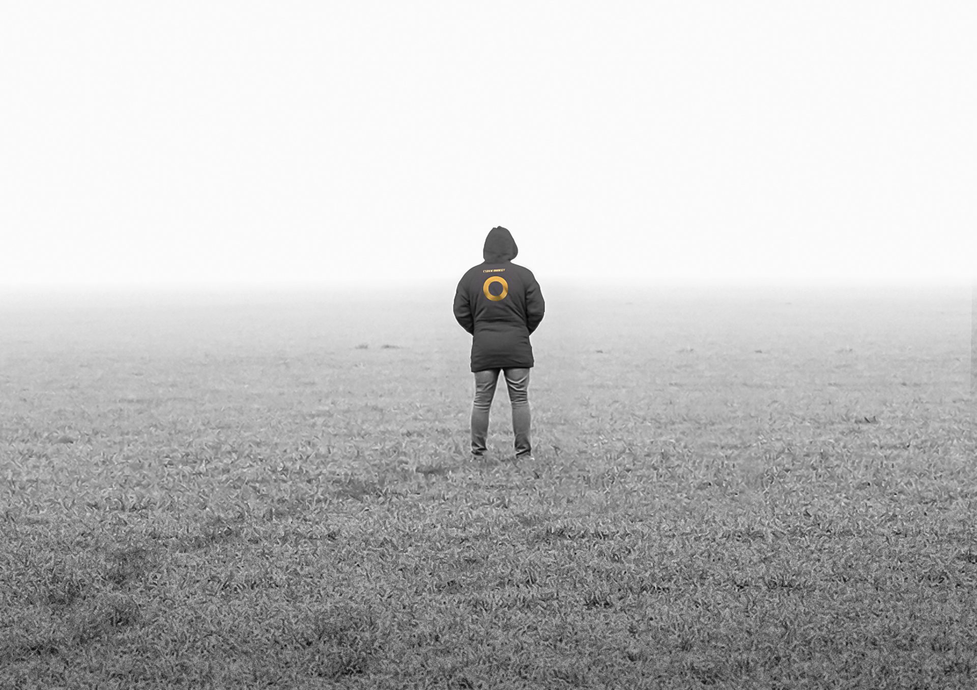 Person in a dark coat with a golden circle logo stands in a vast, misty grayscale field.