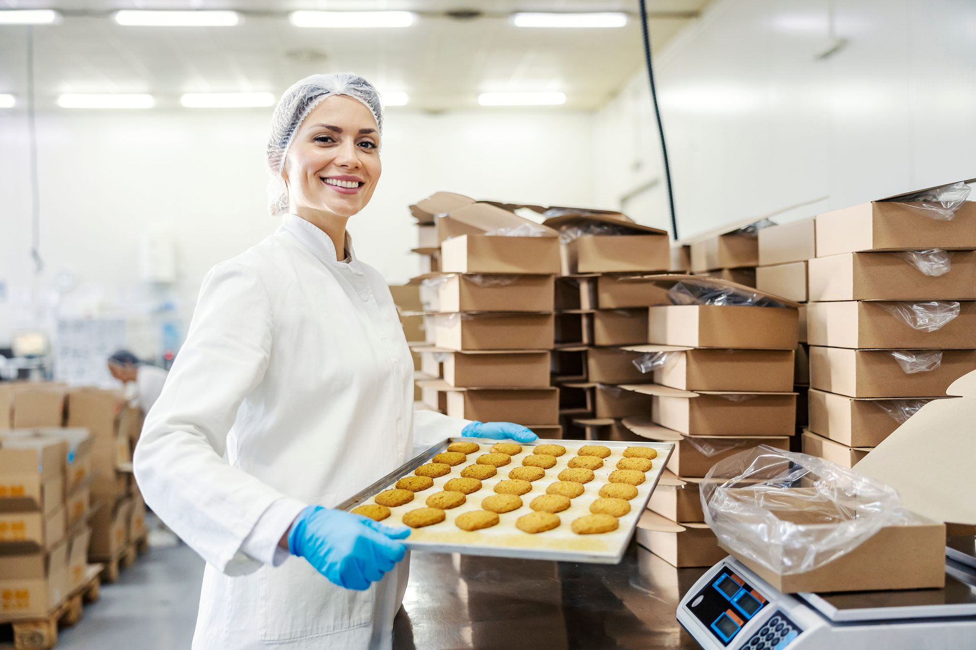 Smiling woman in factory holds cookie tray. Boxes fill background.