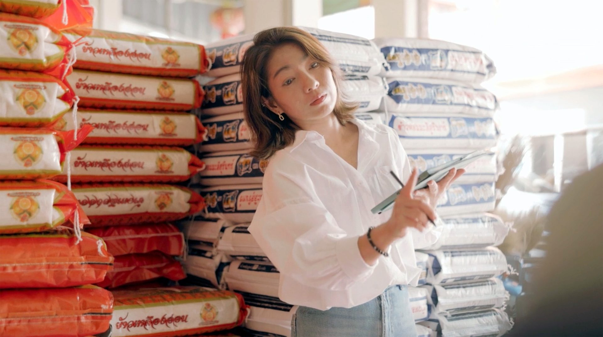 Young woman checking stock on a tablet, surrounded by stacked rice bags in a warehouse.