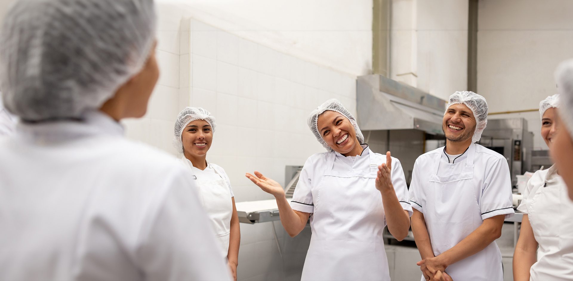 Smiling chefs in hairnets and white coats talking in a commercial kitchen, showing teamwork and joy.