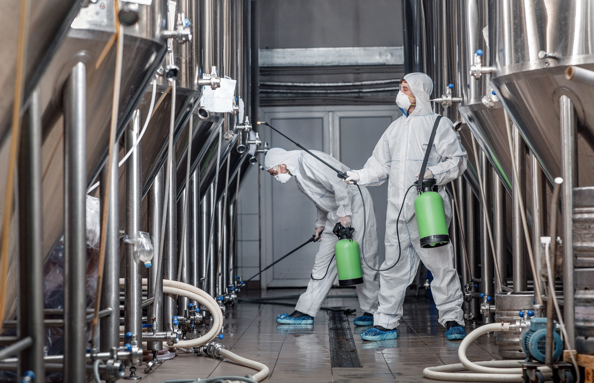 Two masked workers in white protective suits spray large industrial tanks with green sprayers.
