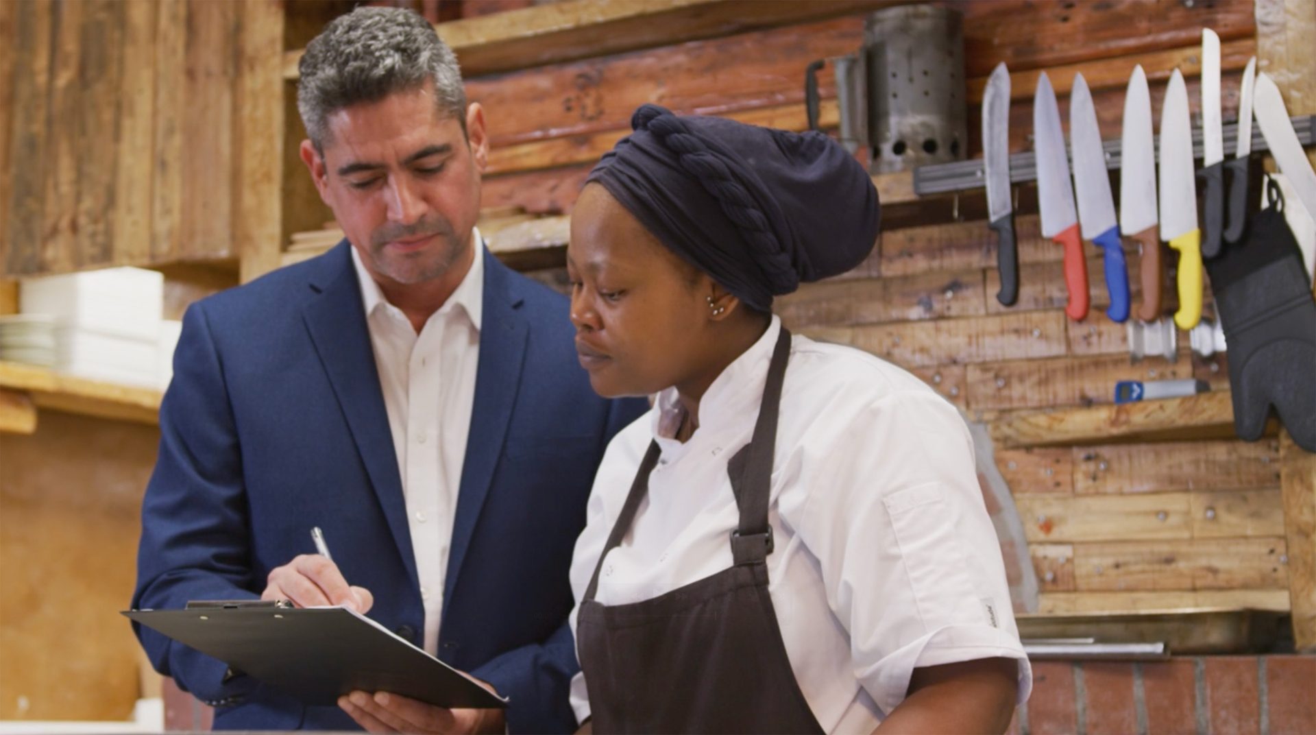 Man in a suit reviews a clipboard with a female chef in a kitchen, possibly discussing restaurant operations.