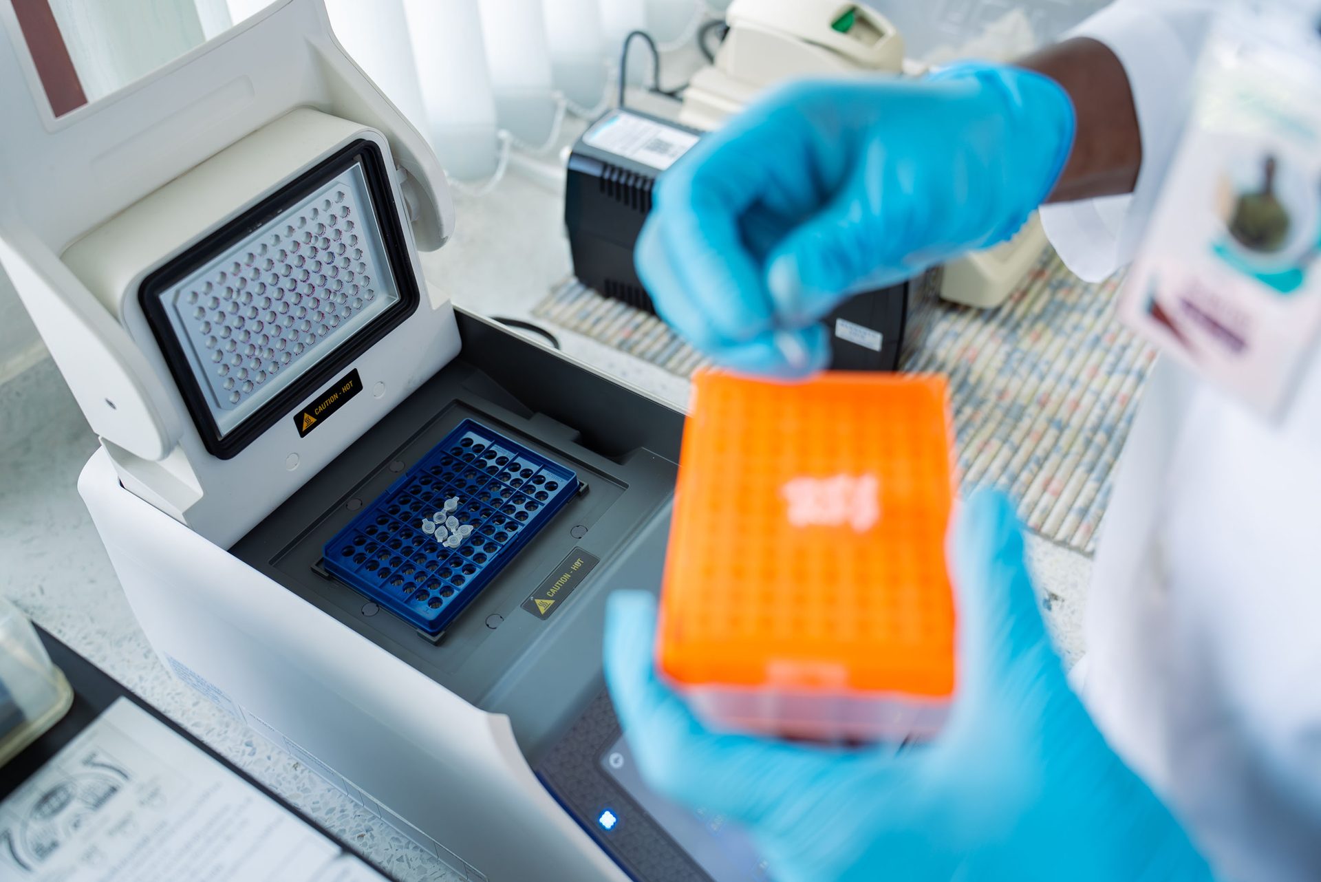 A scientist in blue gloves works with laboratory equipment, including a thermocycler and pipette tips.