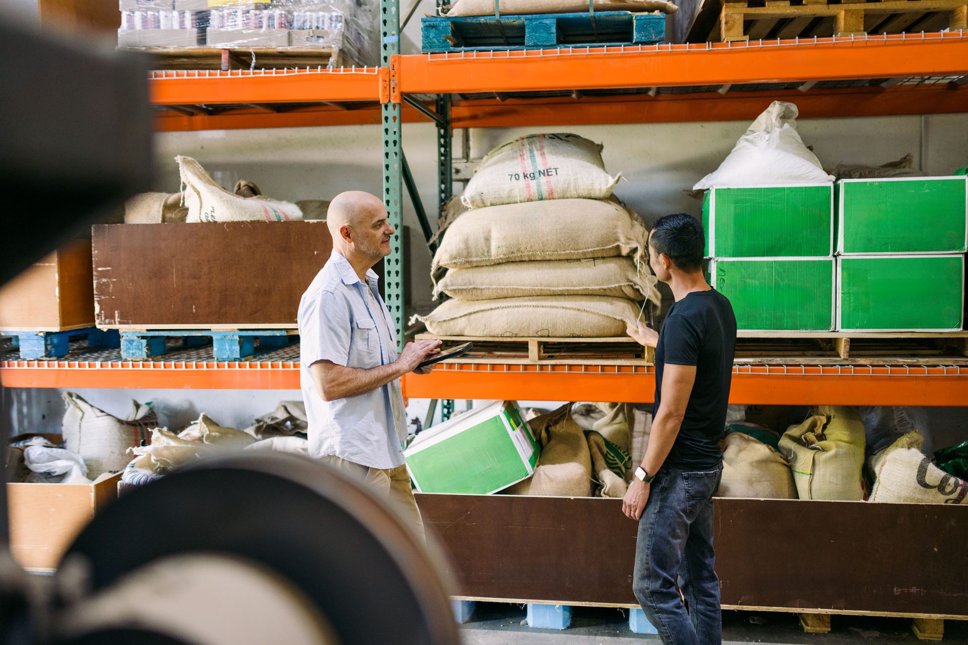 Two men in a warehouse, one with a tablet, inspecting goods on shelves with sacks and boxes.