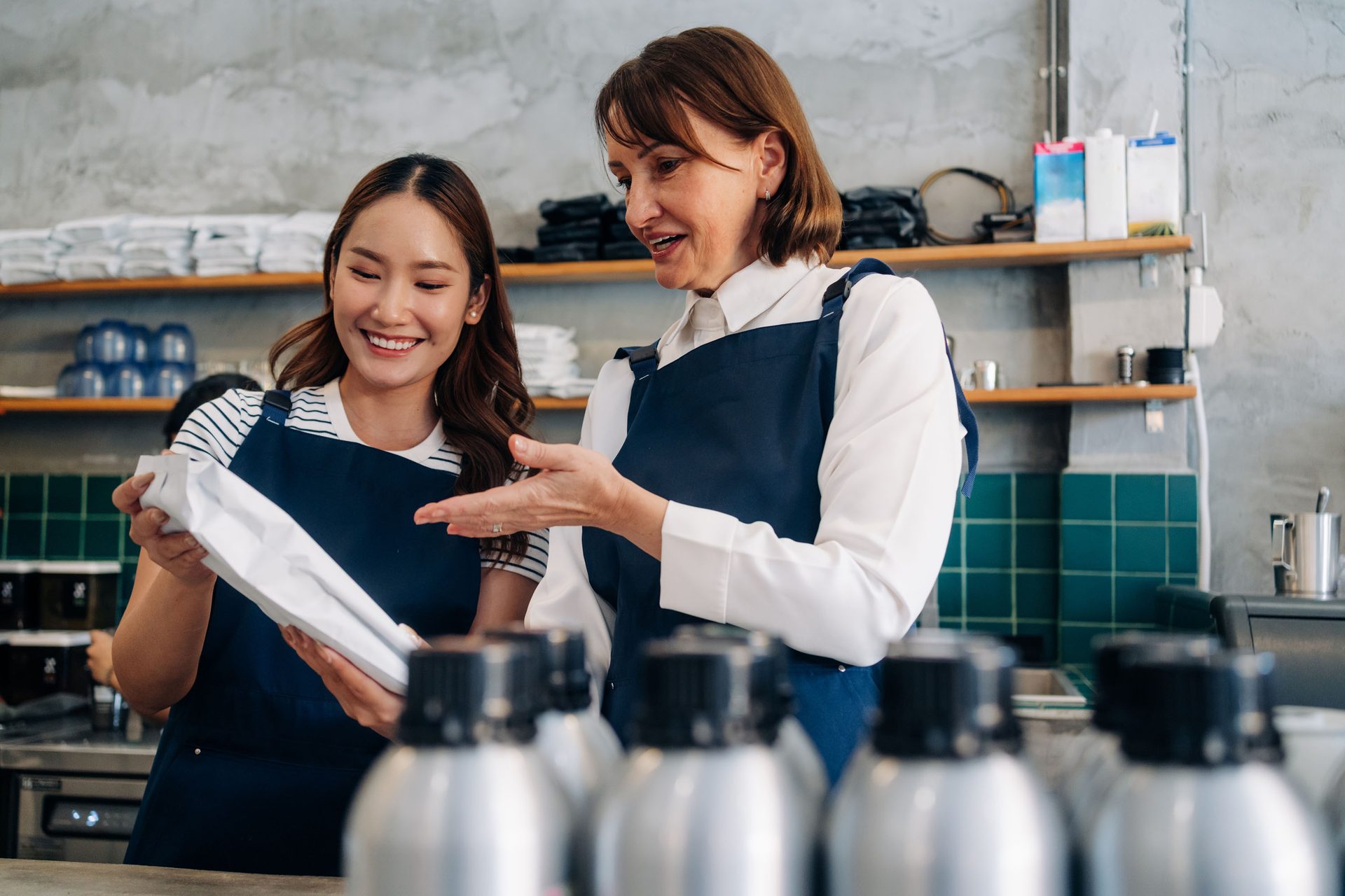 Two smiling baristas, an older woman and a younger woman, discussing a product package in a cafe.