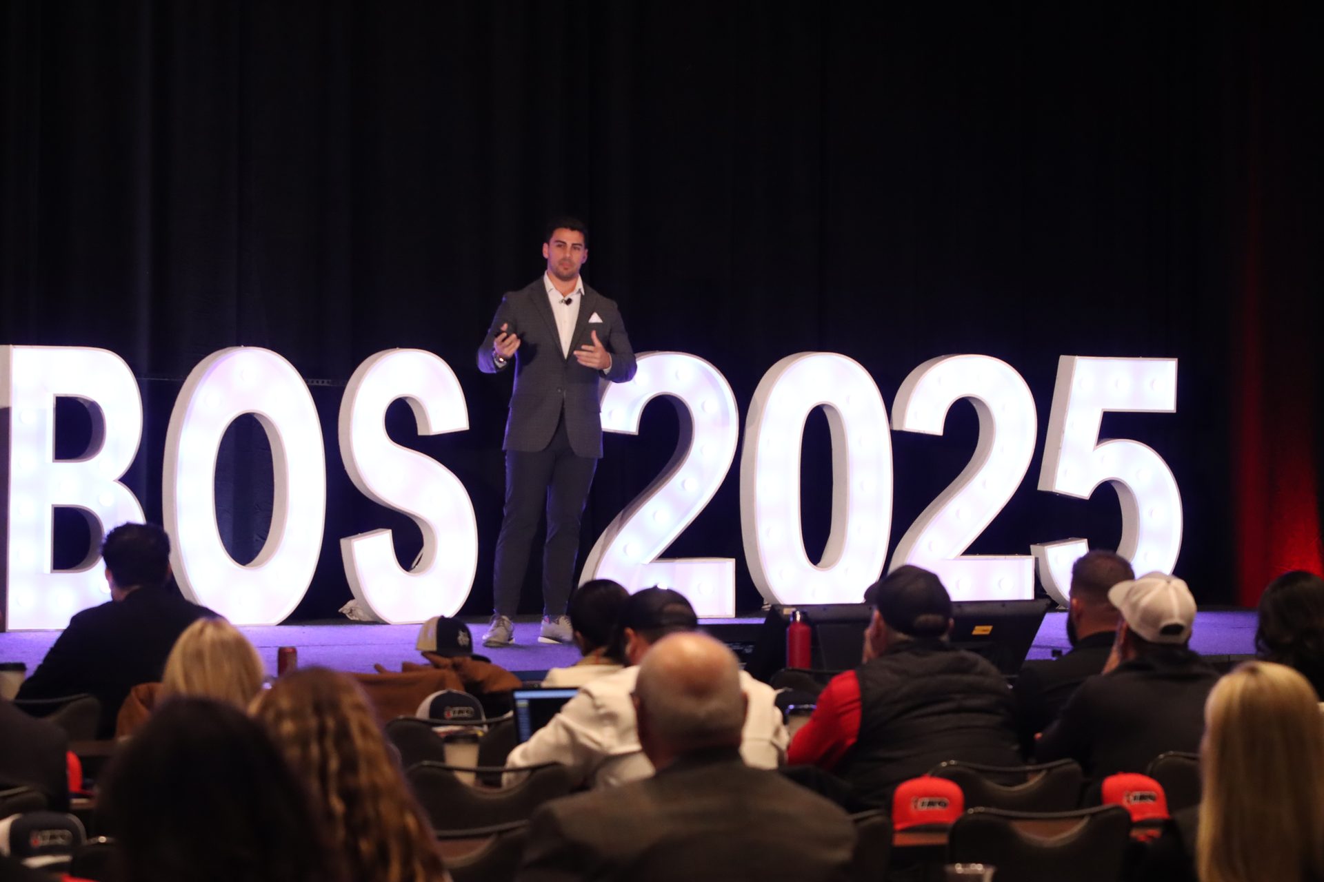 A speaker on stage in front of illuminated "BOS 2025" letters, addressing an audience.