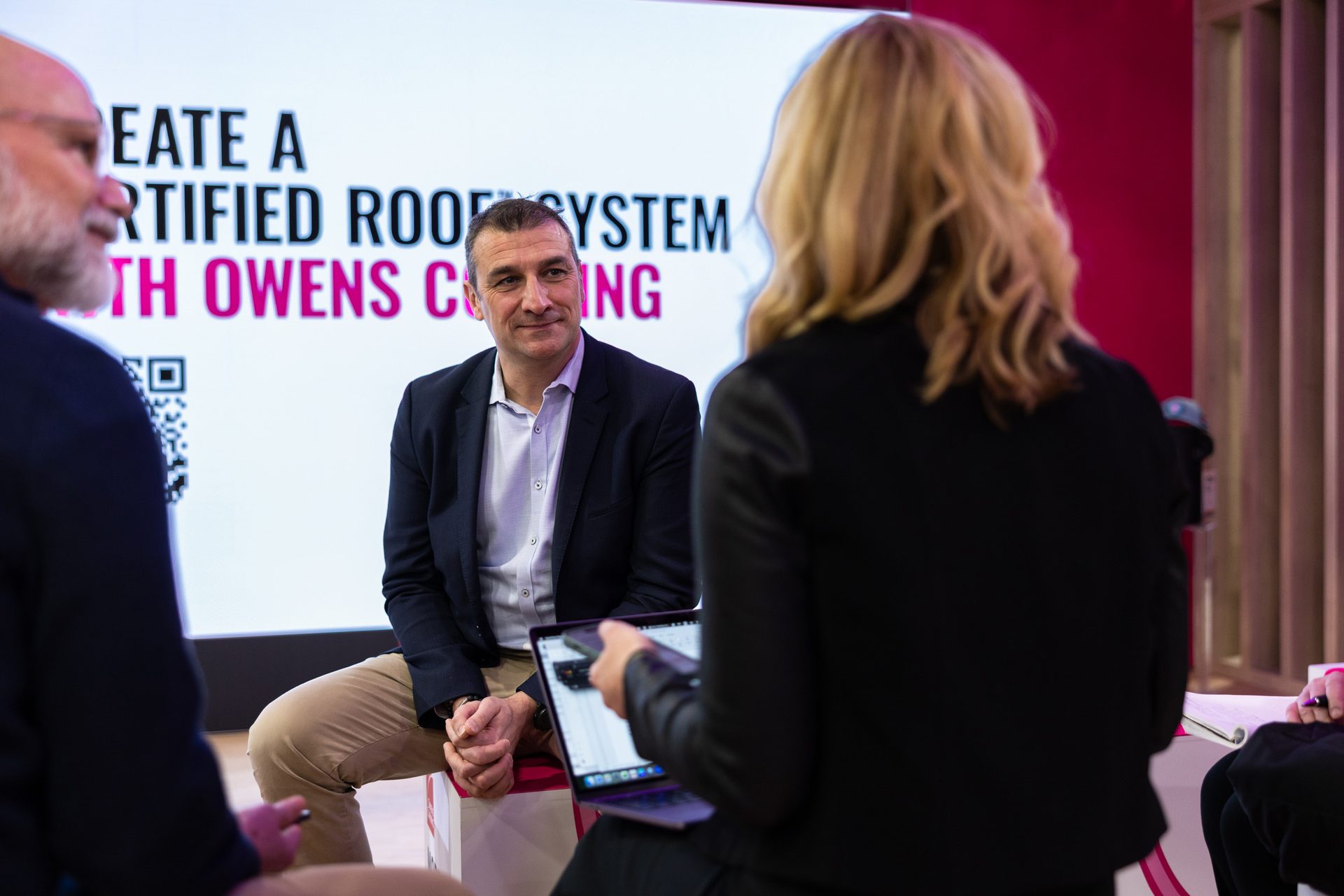 Man in suit converses with two people, possibly an interview, with a 'Certified Roof System' display behind him.