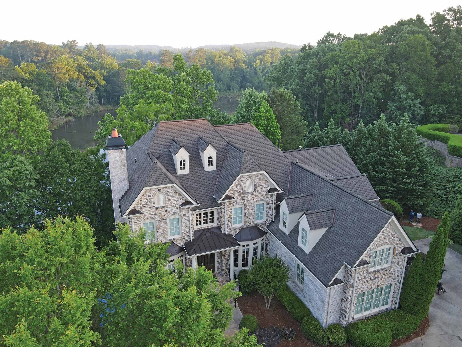 Aerial view of a large stone house with a dark roof, surrounded by trees, overlooking a lake.