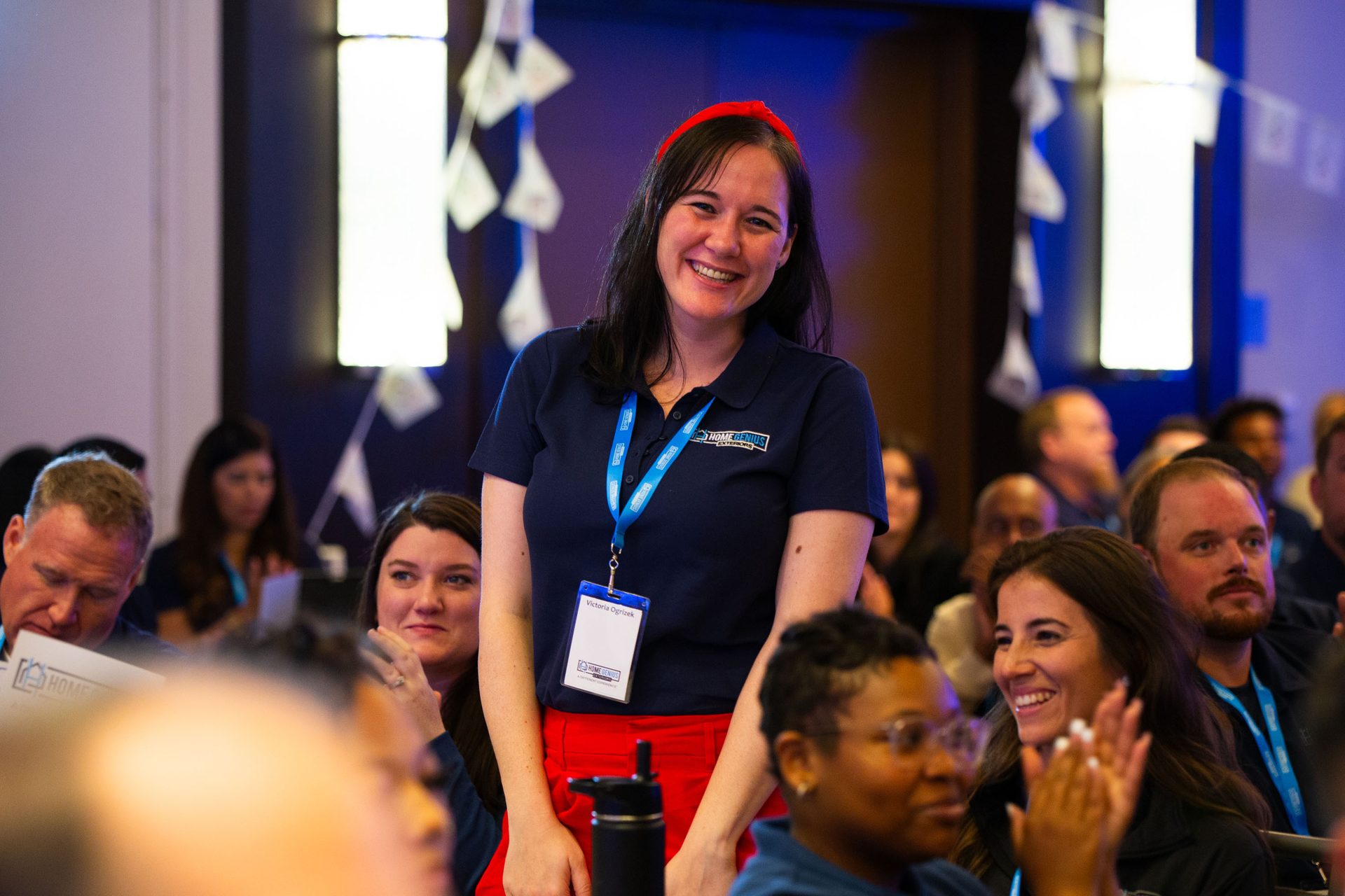 A smiling woman with a red headband stands out in a crowd at an event, wearing a blue polo and red pants.