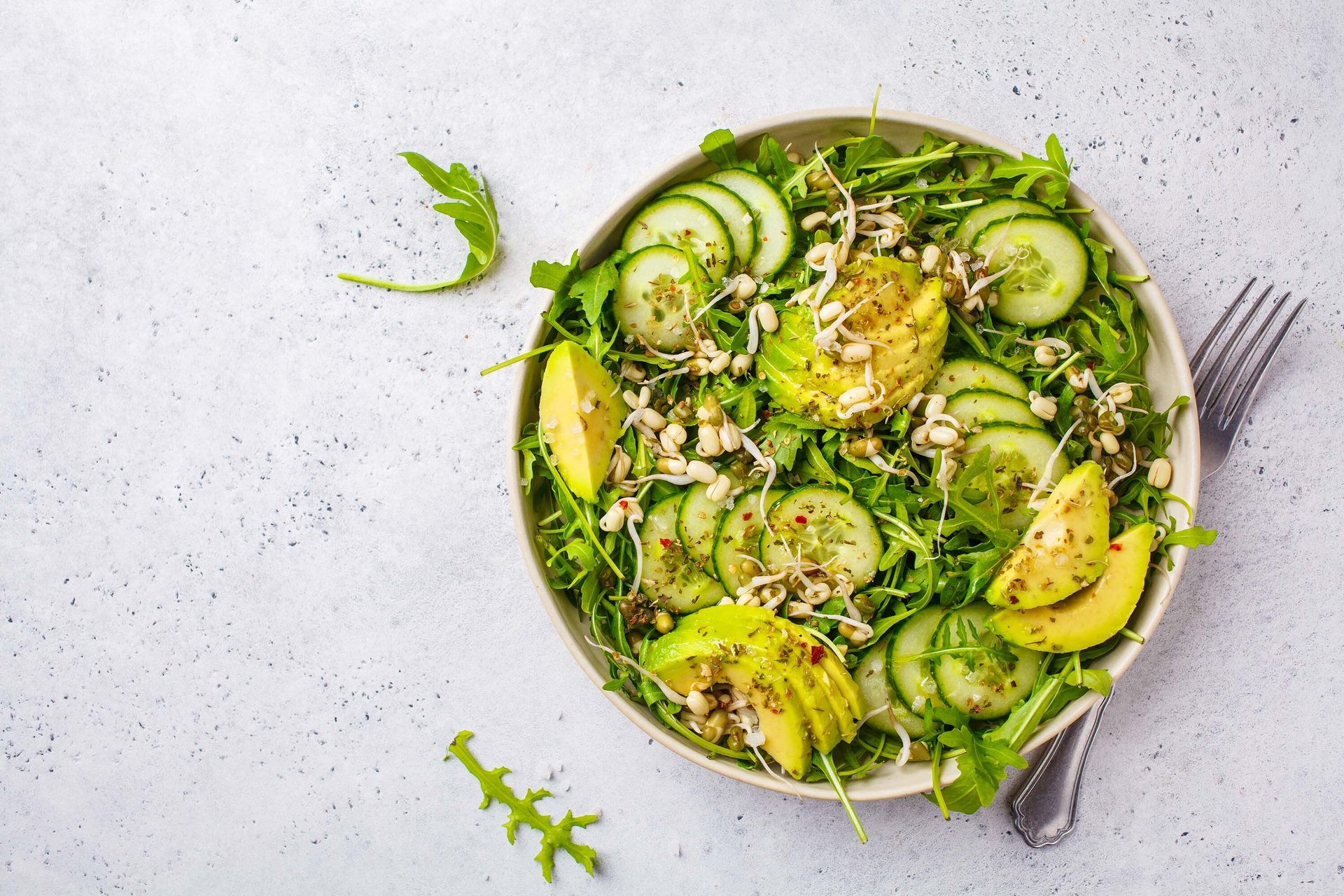 Healthy green salad with arugula, cucumber, avocado, and sprouts in a bowl on a light surface.