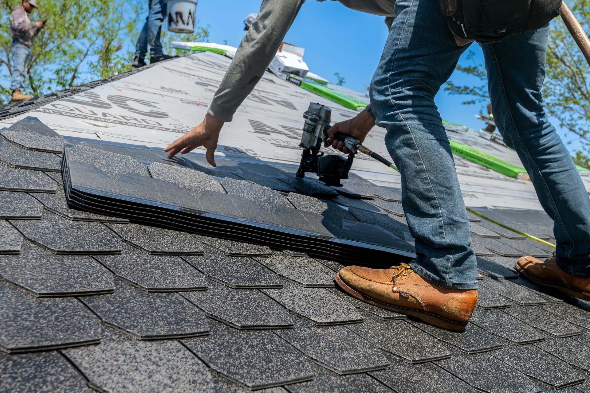 Roofer uses a nail gun to install dark shingles on a roof.