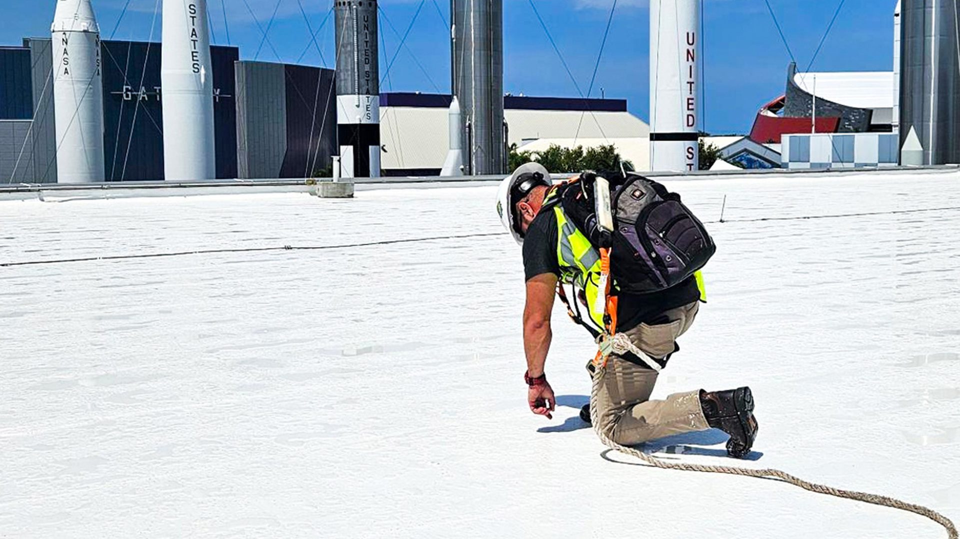 Worker in safety gear kneels on a white rooftop, with rockets and buildings in the background.
