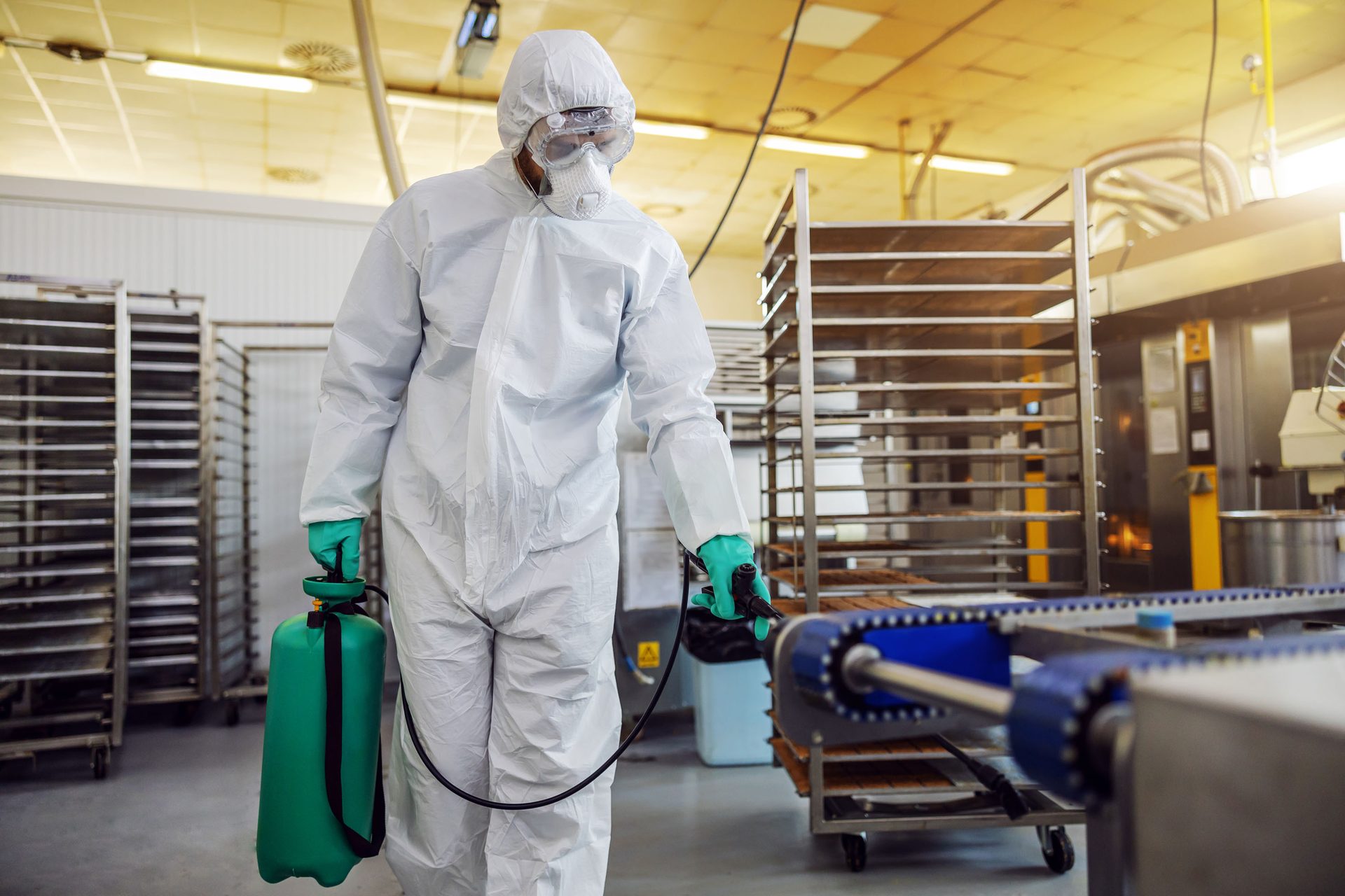 A person in a white hazmat suit, goggles, and respirator sprays disinfectant near a conveyor belt in a factory.