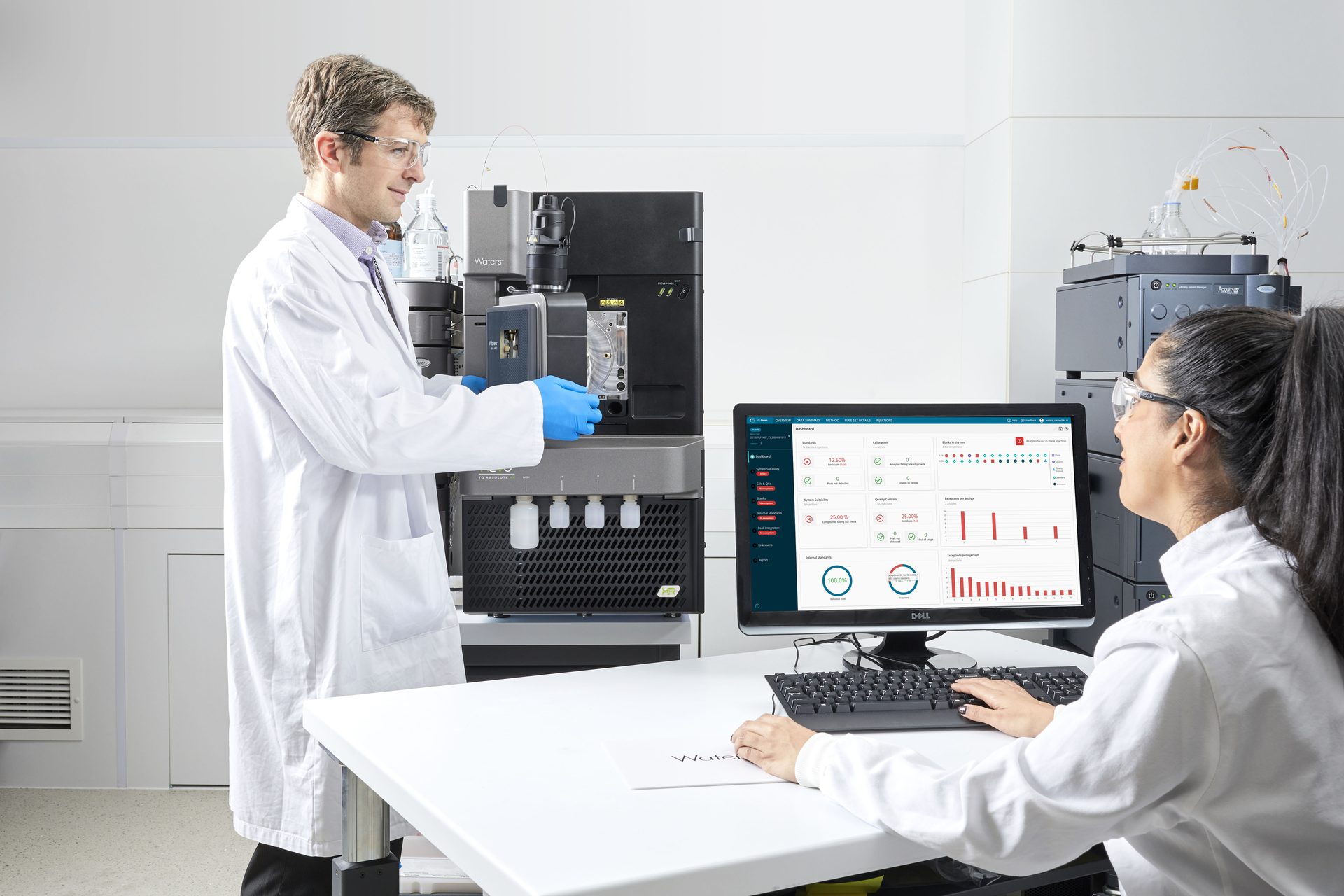 Two scientists, one operating lab equipment and the other monitoring data on a computer screen.