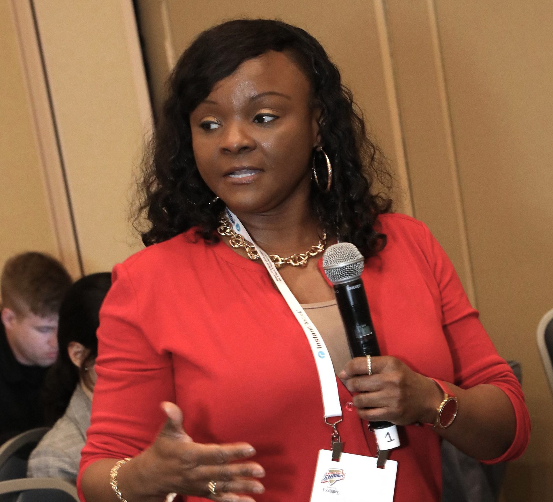 A Black woman in a red top and jewelry speaks into a microphone.