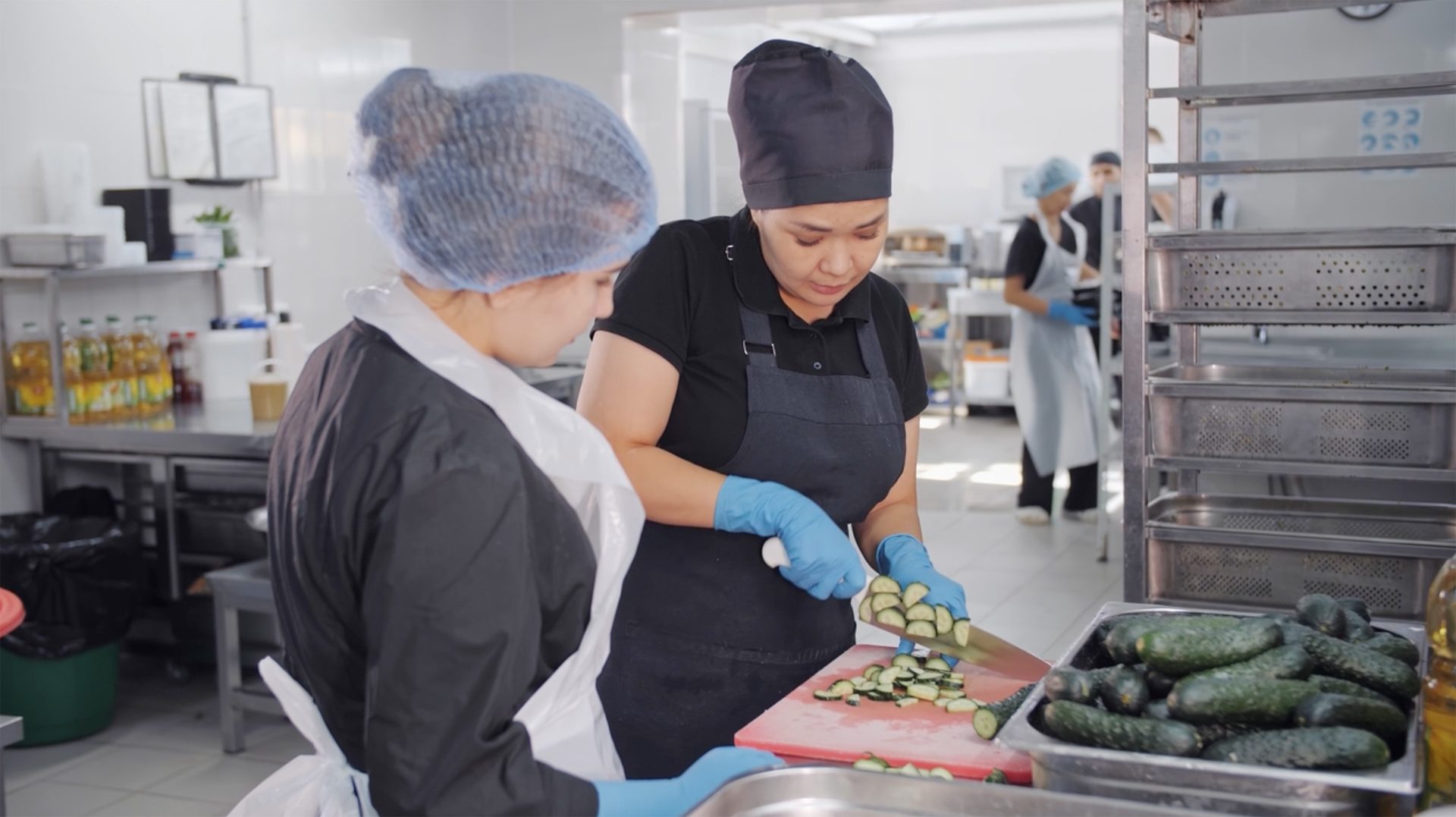 Two chefs in a commercial kitchen preparing food, one chopping cucumbers while the other observes.