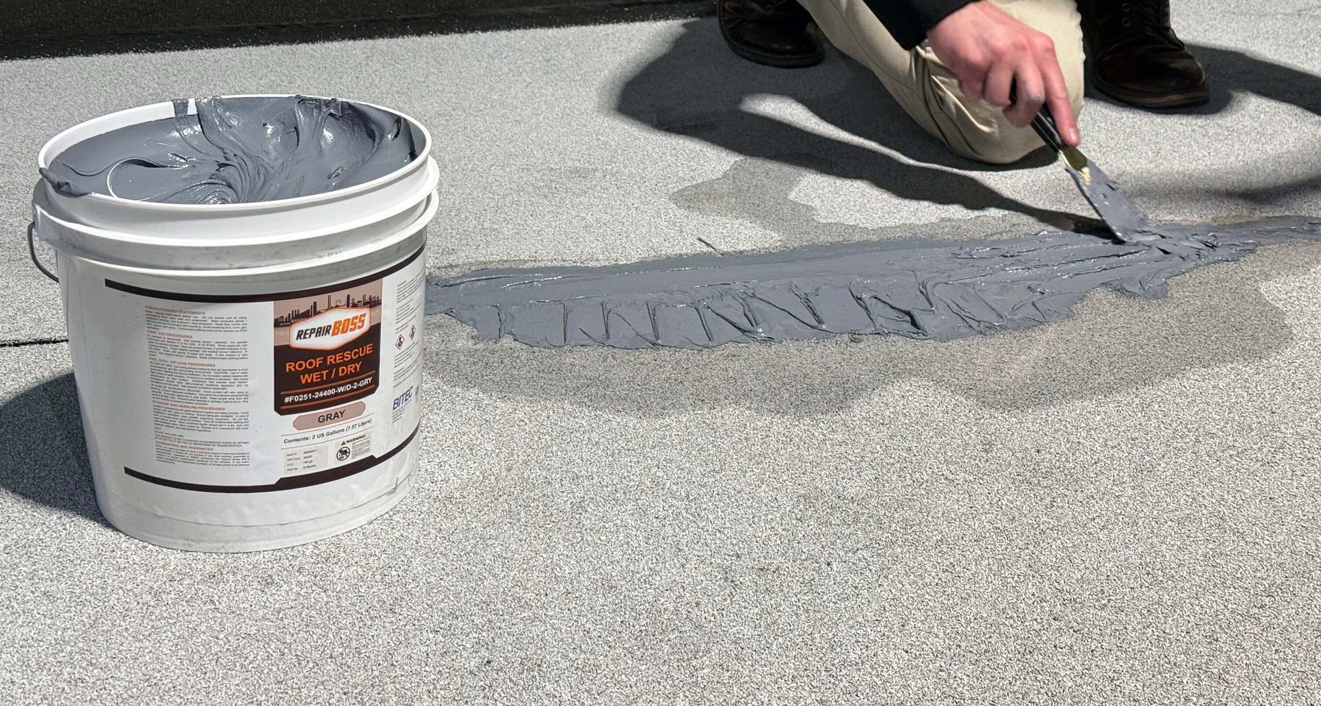Person applies gray roof sealant from a REPAIR BOSS bucket with a trowel onto a granular roof.