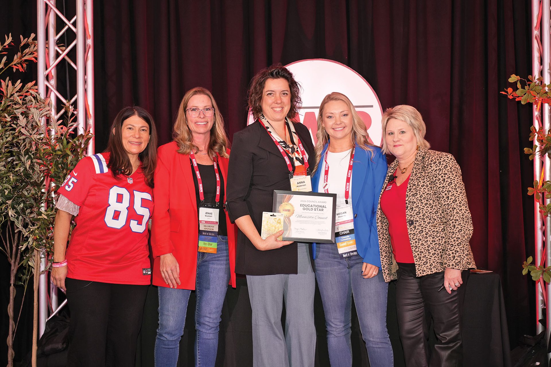 Five smiling women on stage, one holding an award, with a red backdrop and plants on the sides.