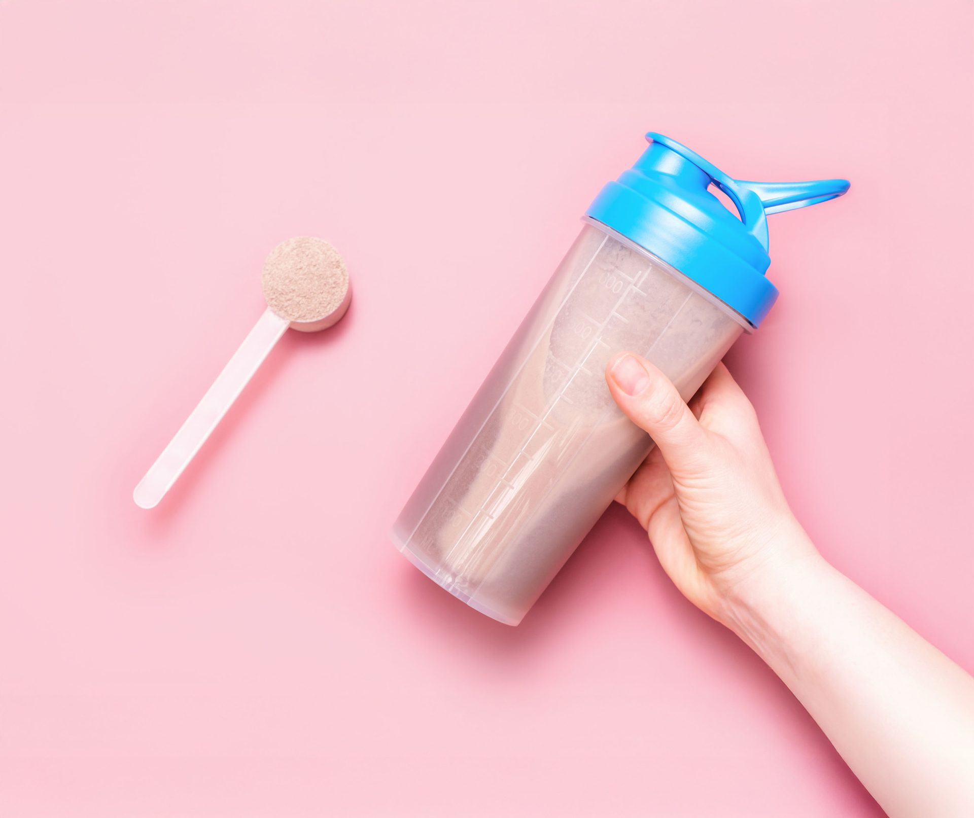 Hand holding a protein shaker and a scoop of protein powder on a pink background.