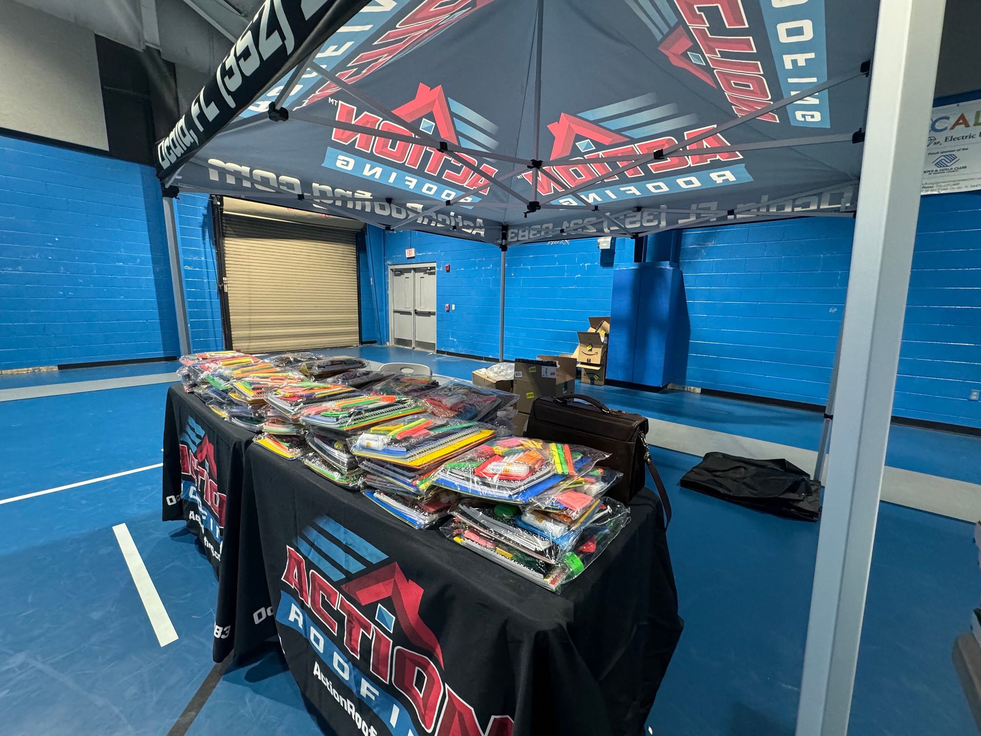 ACTION ROOFING tent over table with packaged items in a blue gym.