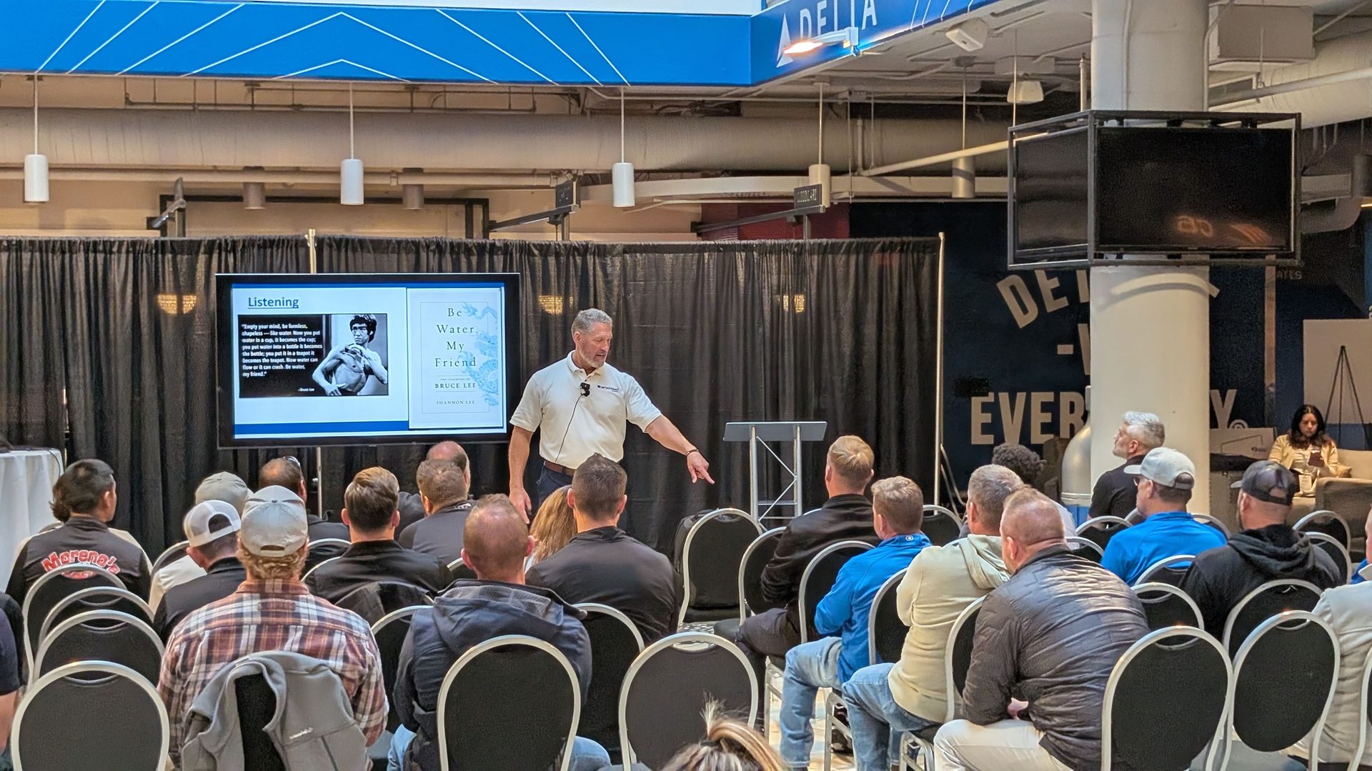 Man speaks to an audience, pointing at a screen with Bruce Lee's 'Be Water, My Friend' quote.