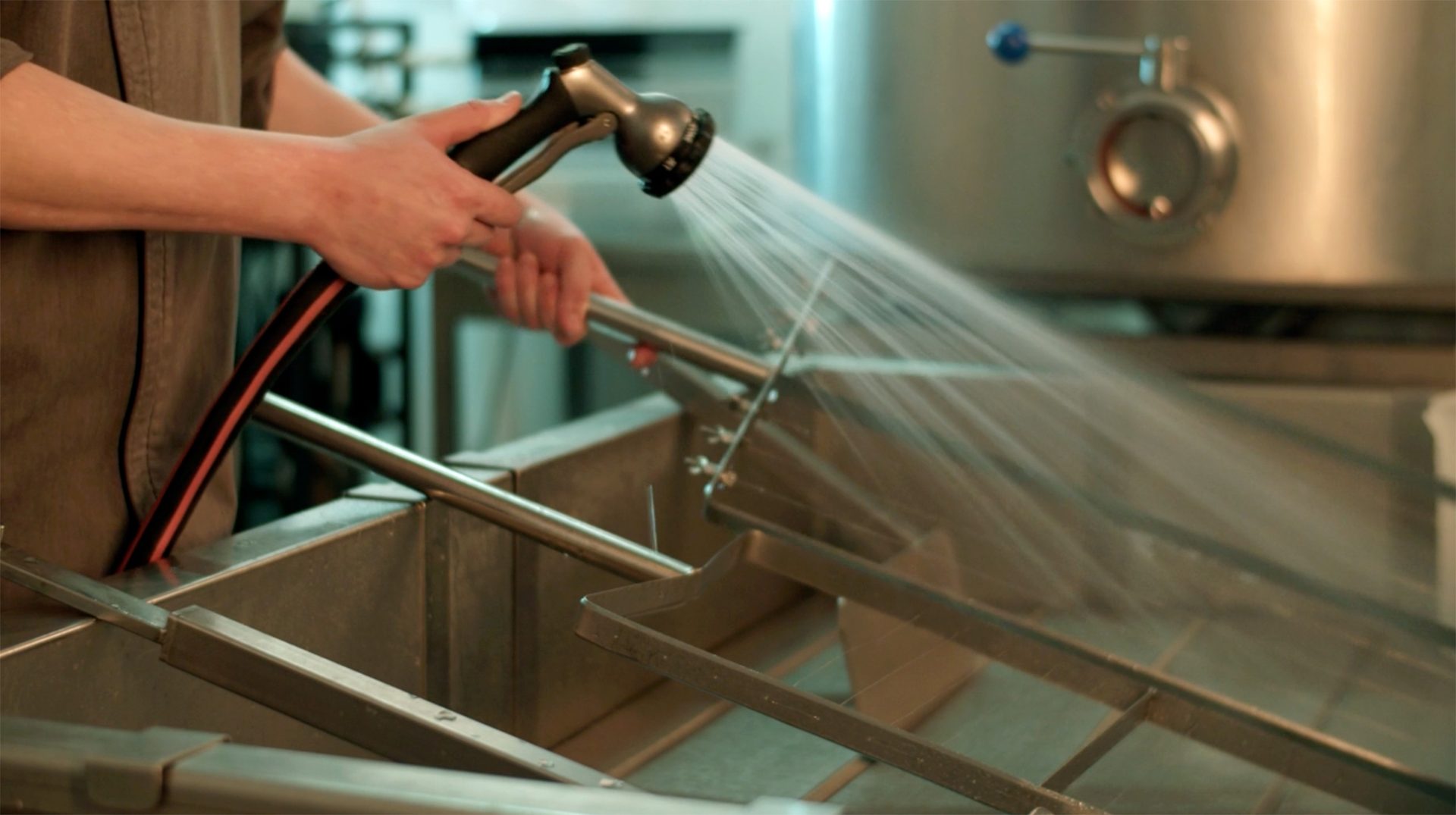 Person's hands holding a spray nozzle, washing metal equipment with water in a commercial kitchen.