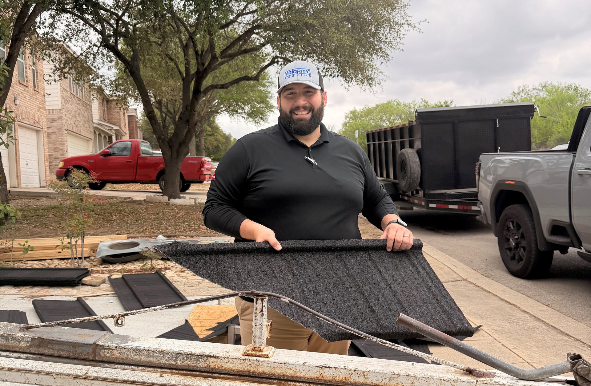 A man in a Priority Roofing hat holds a black metal shingle sample, smiling, with roofing materials nearby.