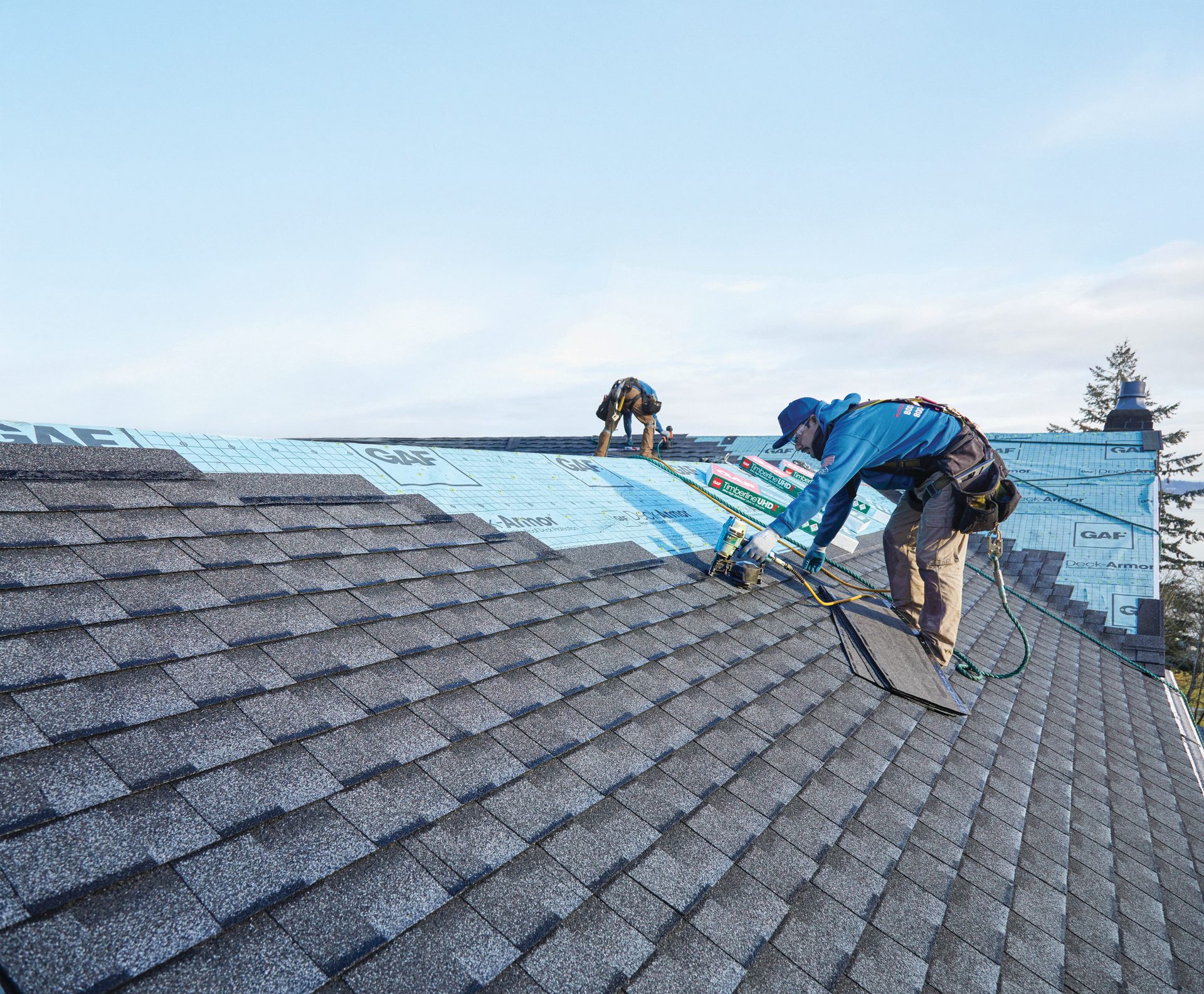 Roofers install dark shingles on a roof covered with blue GAF underlayment, wearing safety harnesses.