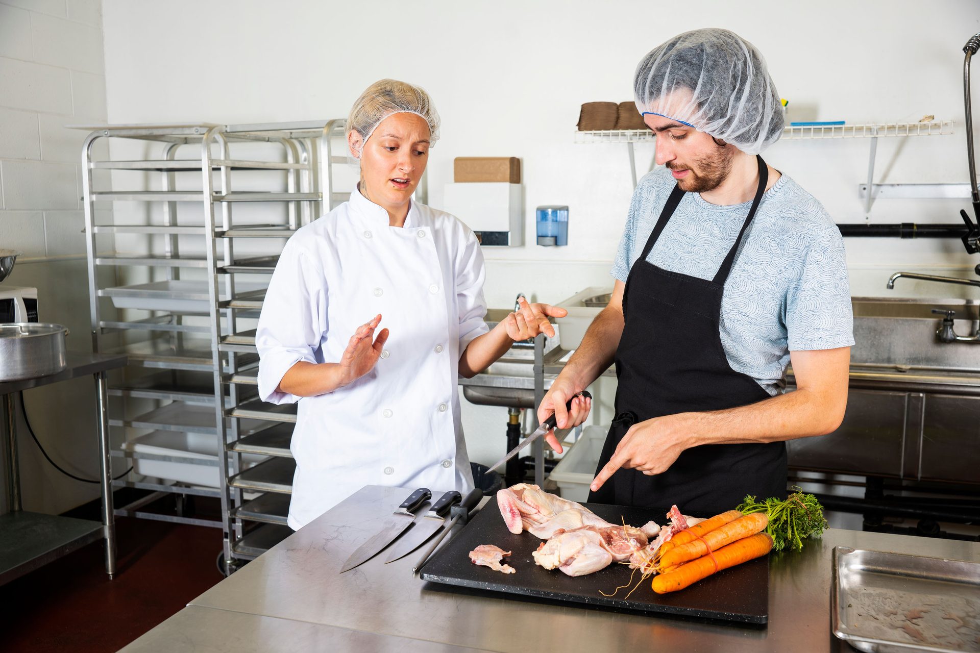 Two chefs, one female and one male, preparing raw chicken and carrots in a professional kitchen.