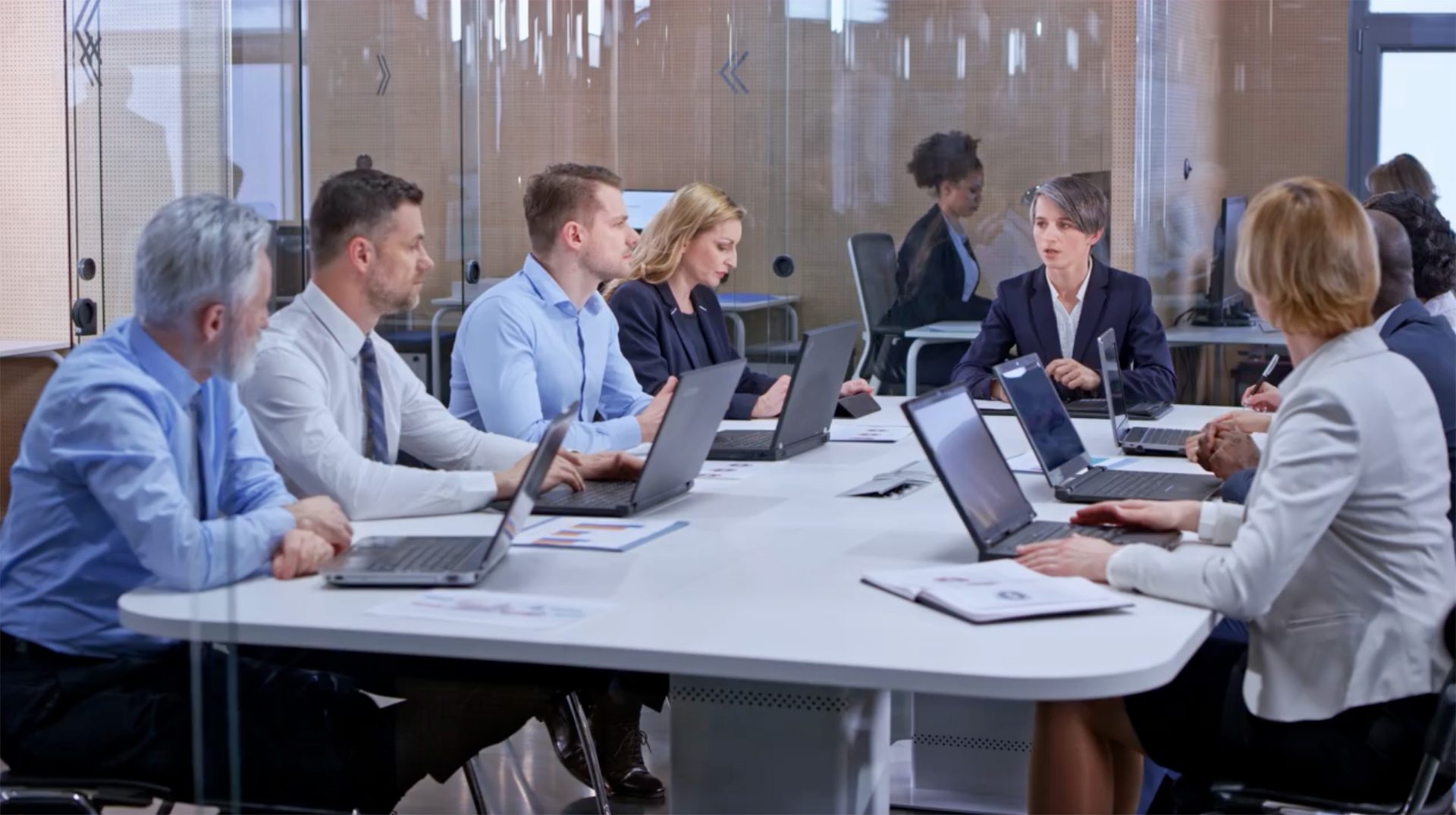 Business professionals in a modern meeting room, engaged in a discussion around a table with laptops.