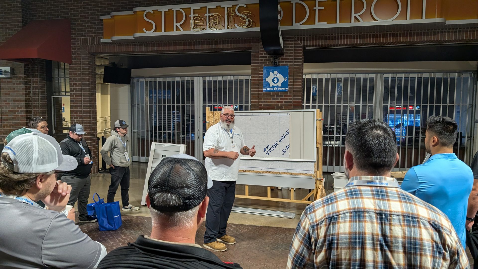 A bearded man instructs a group of people in front of a building display board, "STREETS DETROIT" visible.
