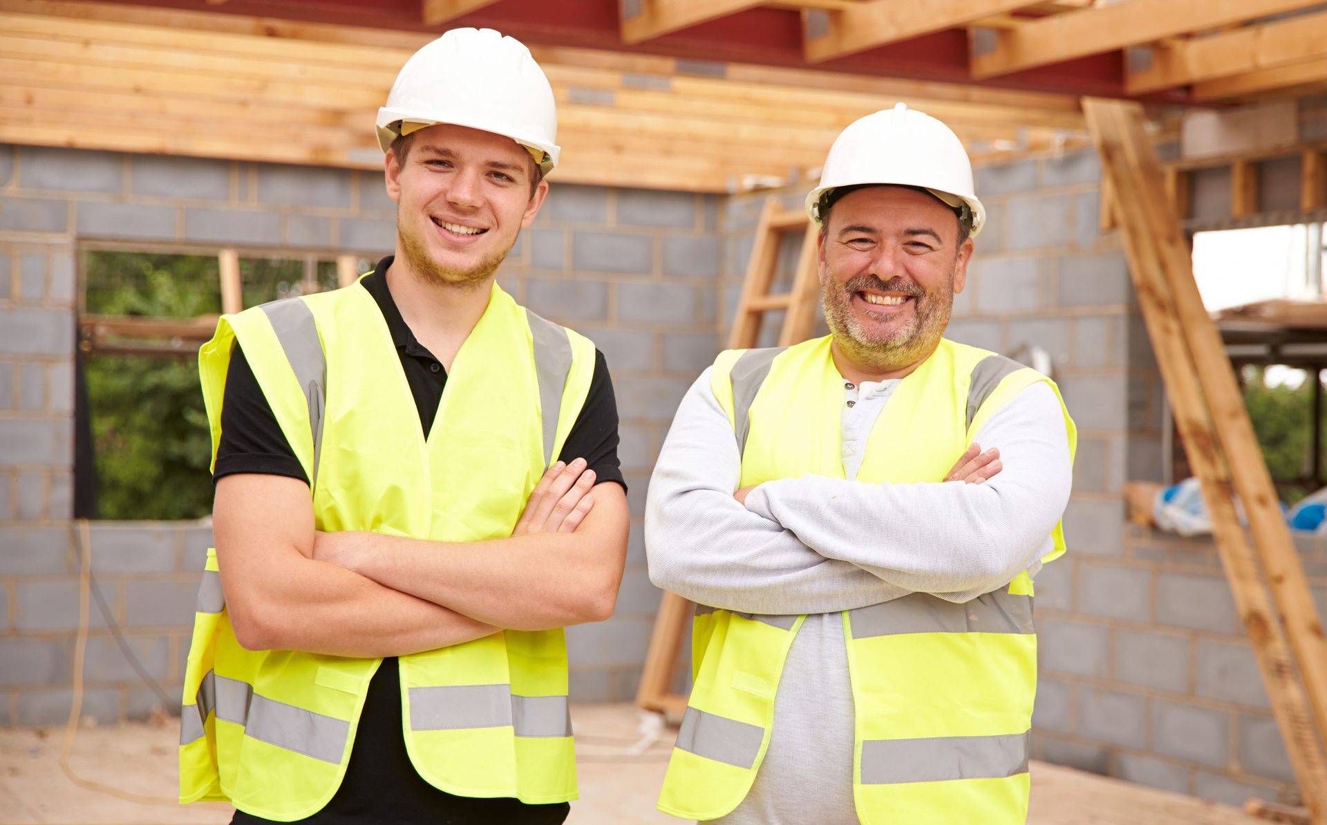 Two smiling construction workers in hard hats and high-vis vests stand with crossed arms at a building site.