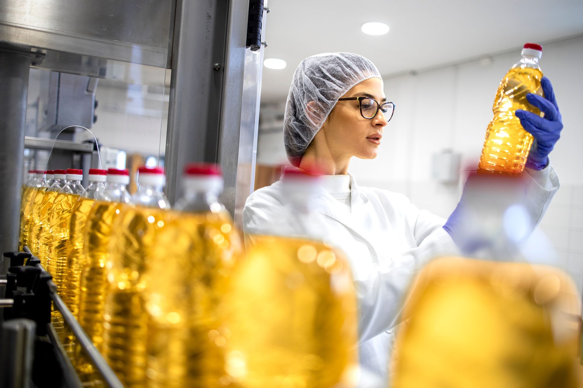 A quality control worker in a hairnet and lab coat inspects a bottle of cooking oil on a factory production line.