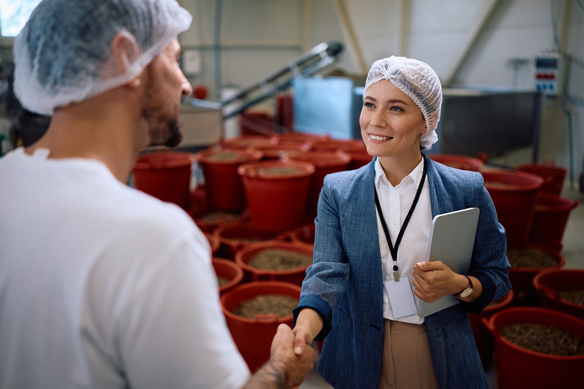 Two people in hairnets shaking hands in a processing plant; woman smiling, holding tablet.