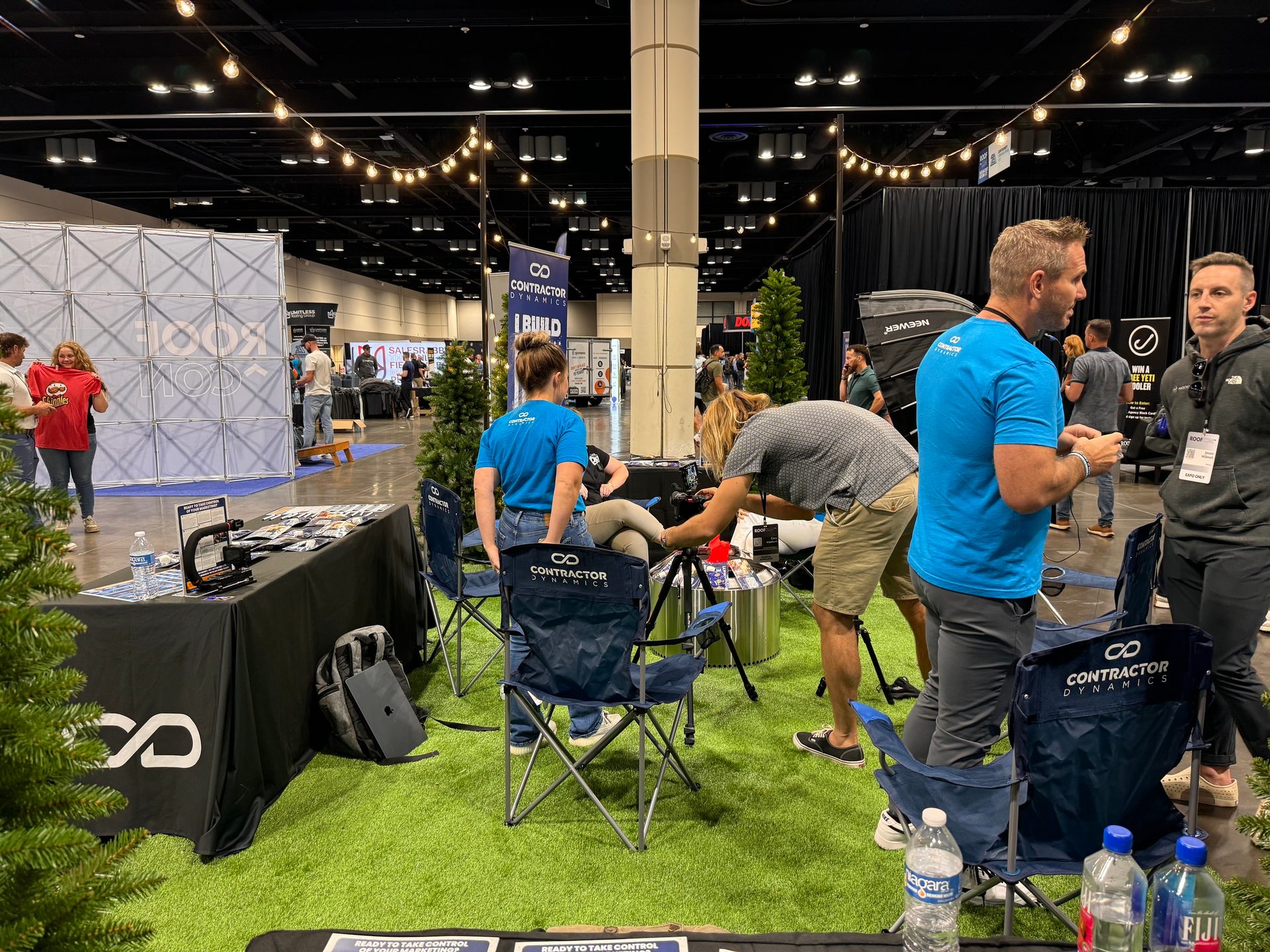 People at an indoor trade show booth with artificial grass, featuring "Contractor Dynamics" branding.