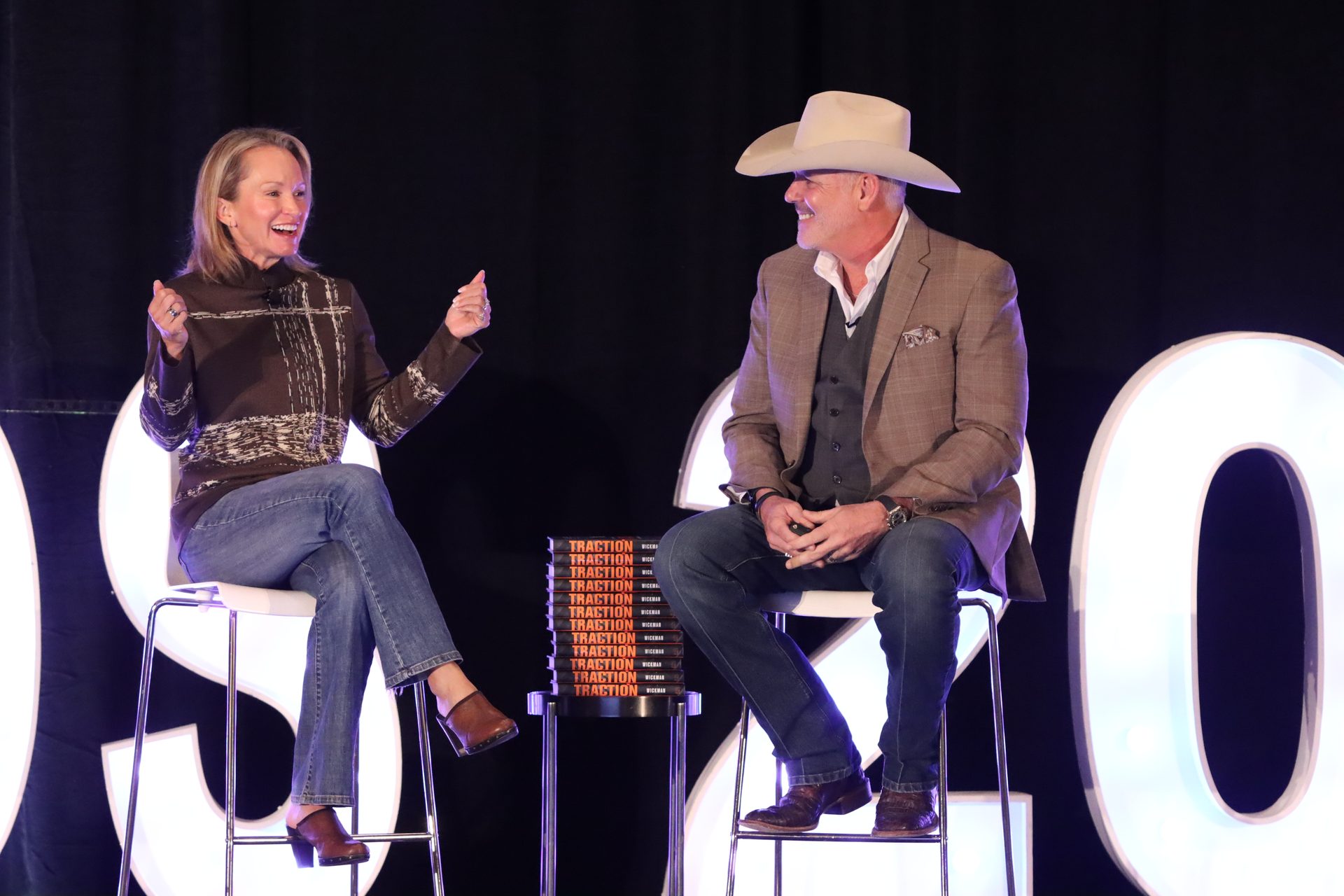 Two people, one in a cowboy hat, smiling and talking on a stage with a stack of "Traction" books.