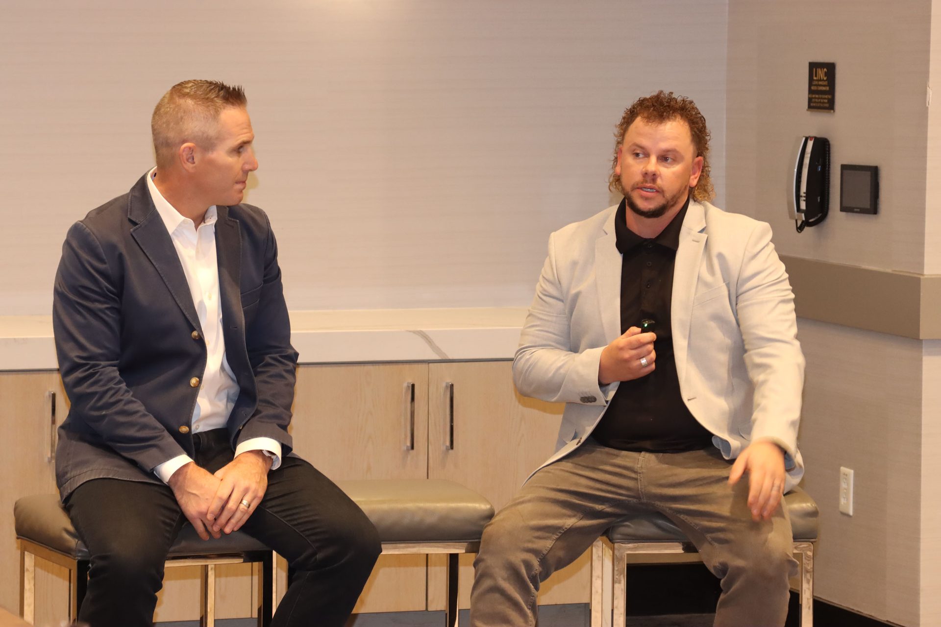 Two men in blazers seated on stools, one with short hair, the other with curly hair, in conversation.