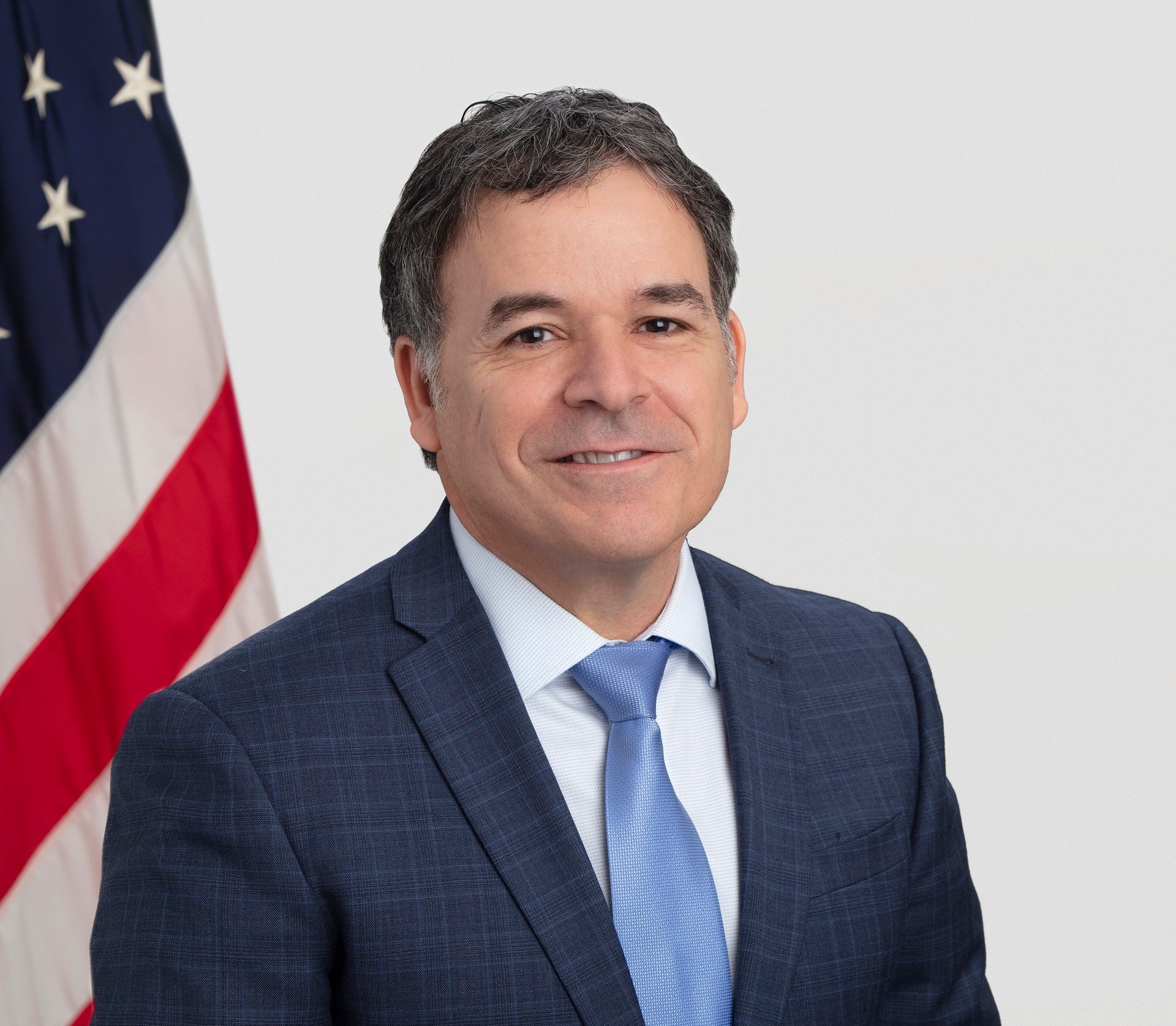 Smiling man in a blue checkered suit and light blue tie, with an American flag backdrop.
