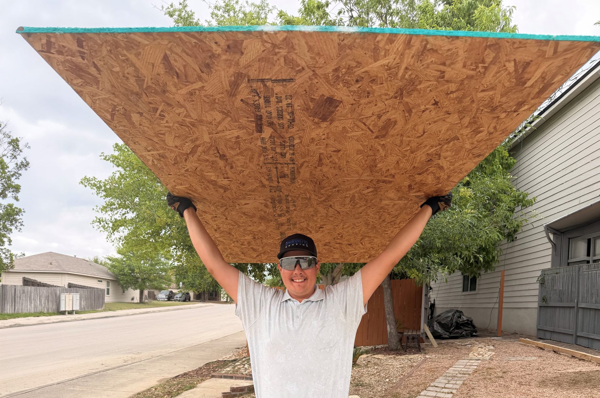 A man in sunglasses and a cap smiles at the camera while holding a large sheet of OSB plywood above his head.