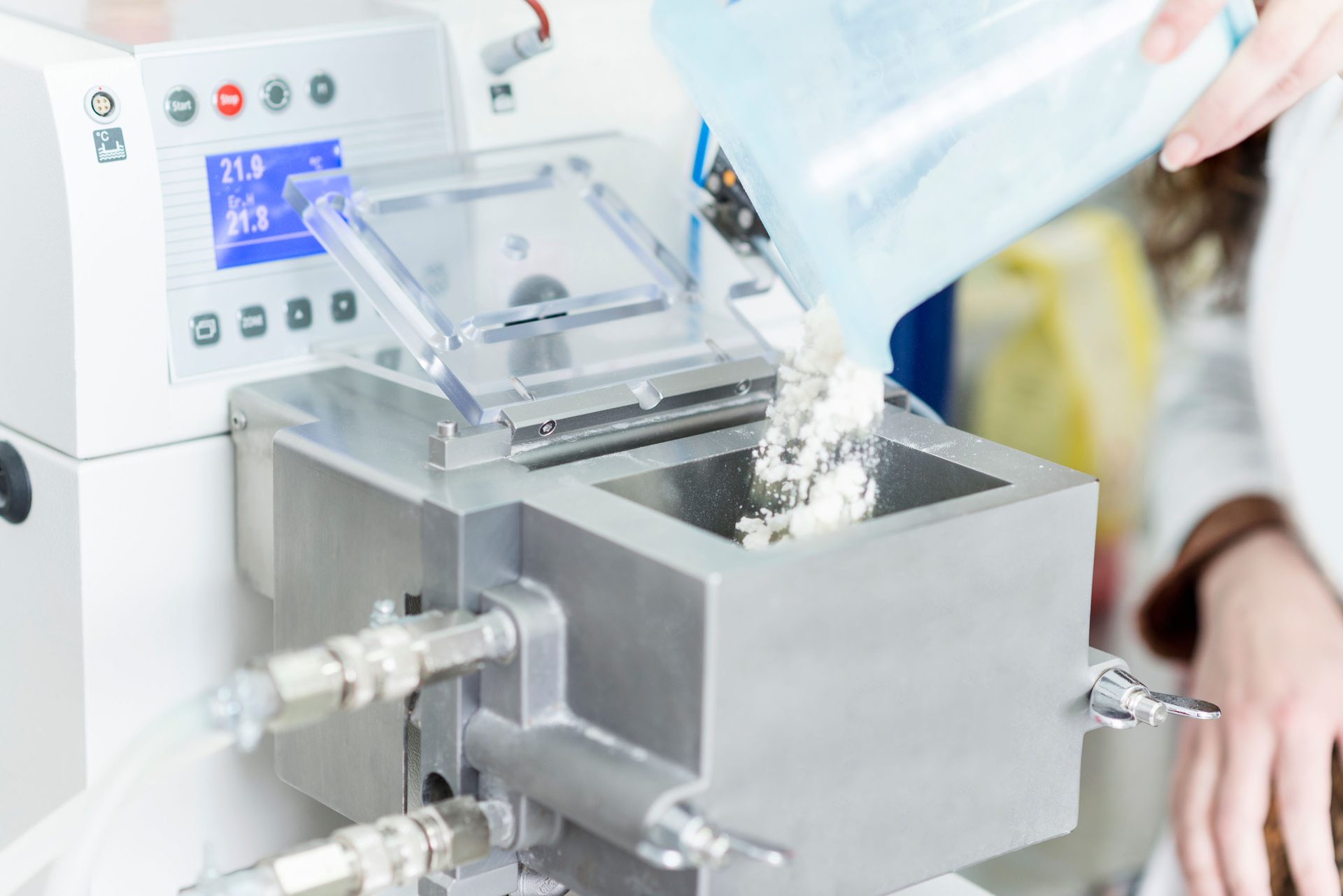 A person pours white powder from a container into a laboratory machine with a digital display.