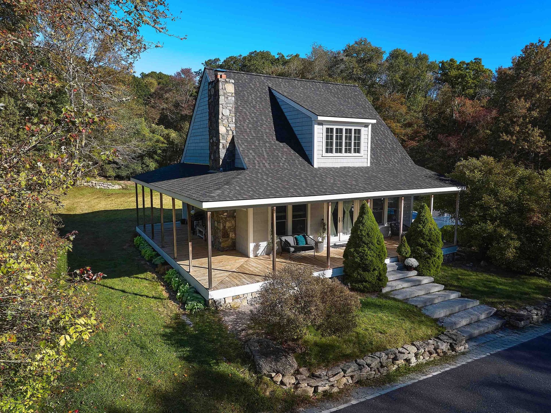 A cozy house with a wraparound porch, stone chimney, and dormer window, set among autumn trees.