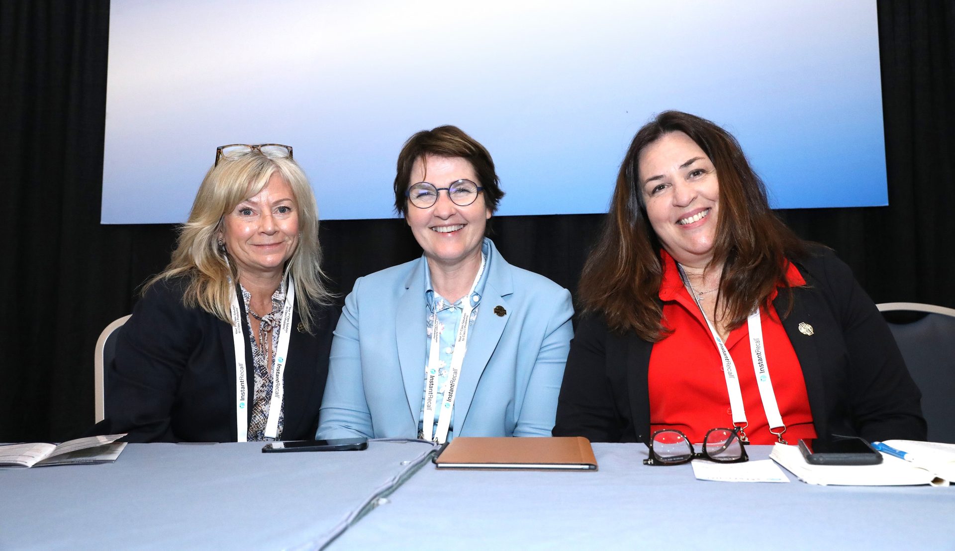 Three smiling women sit at a conference table, wearing lanyards.