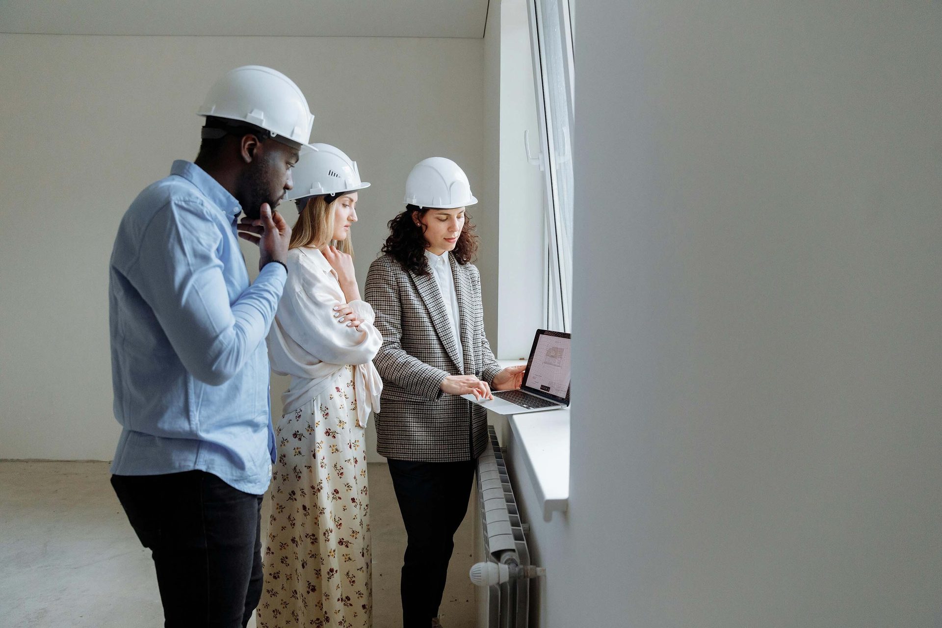 Three people in hard hats review plans on a laptop by a window.