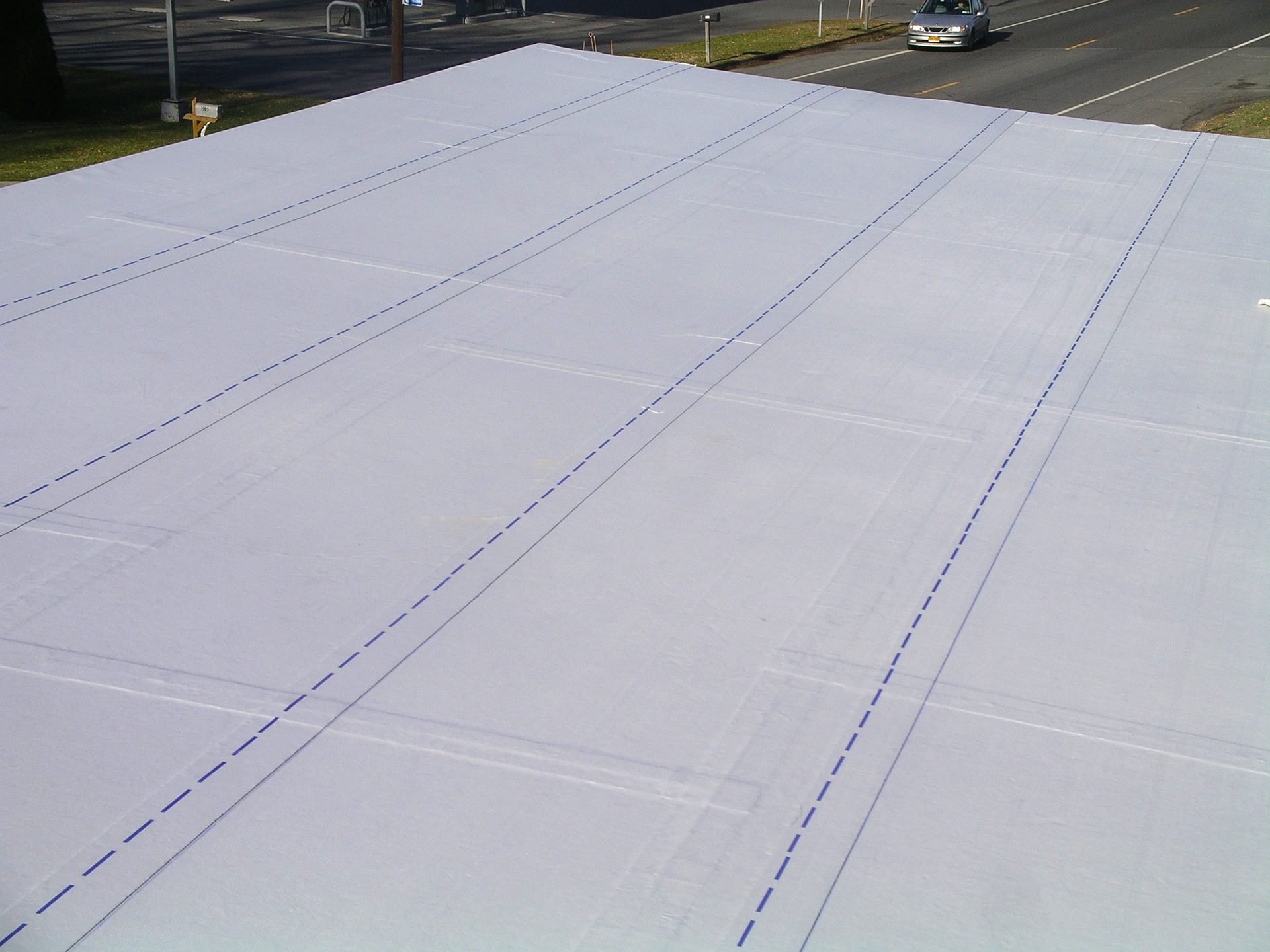 Overhead view of a large white tarp or roof with blue dashed lines. A road and car are in the background.
